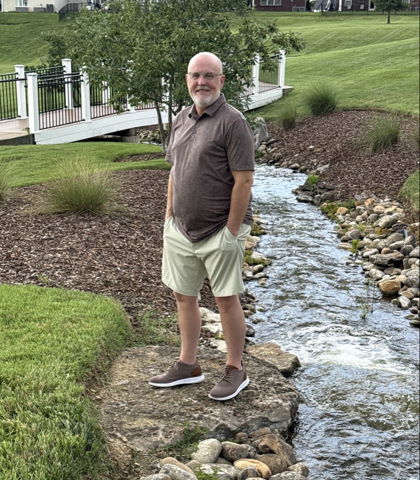 A man standing on a rock by a small creek in a park with neatly maintained grass, trees, plants, and a white bridge in the background.