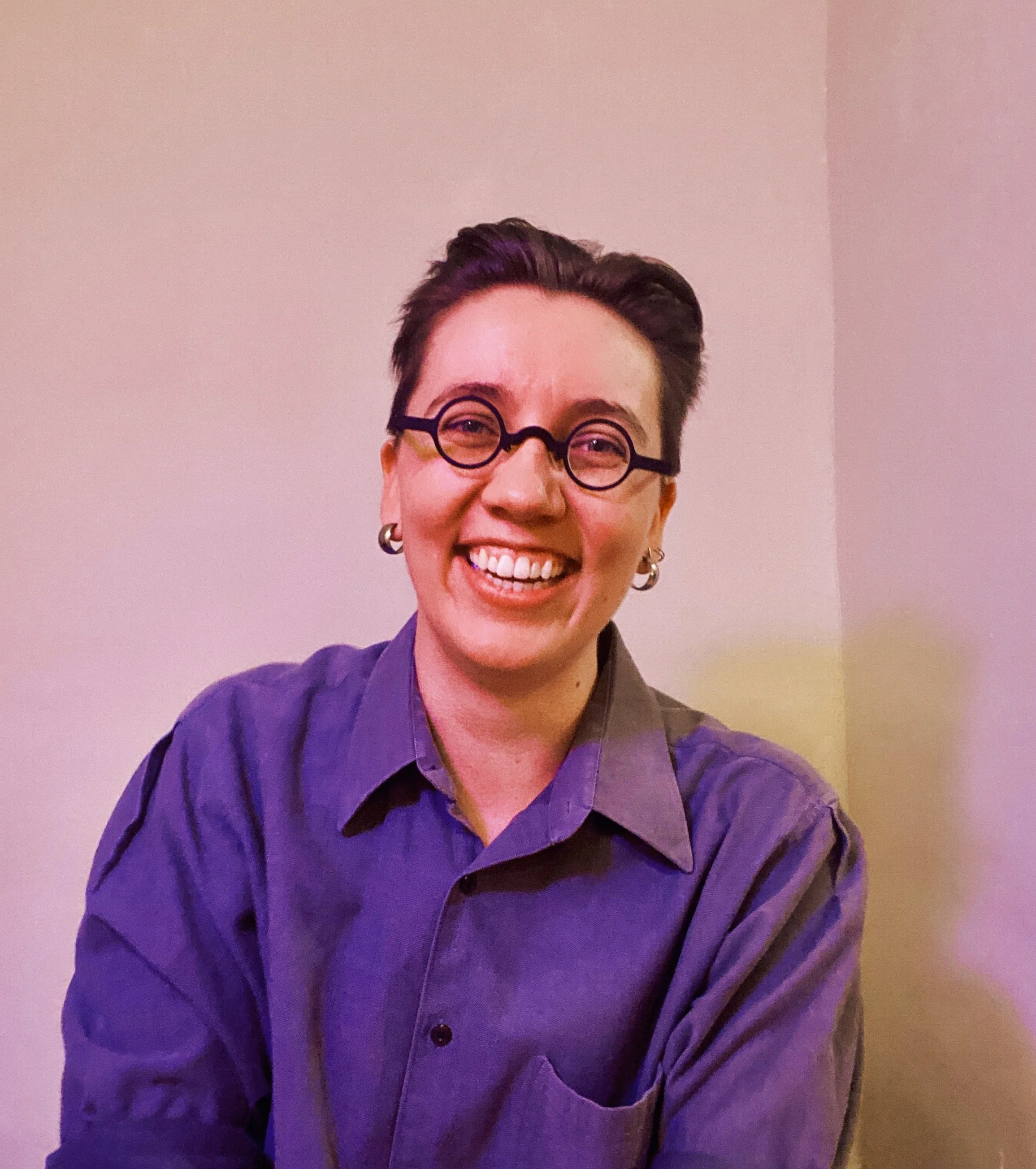 A person with short dark hair, wearing round black glasses and a dark purple shirt, smiling broadly in front of a plain wall.