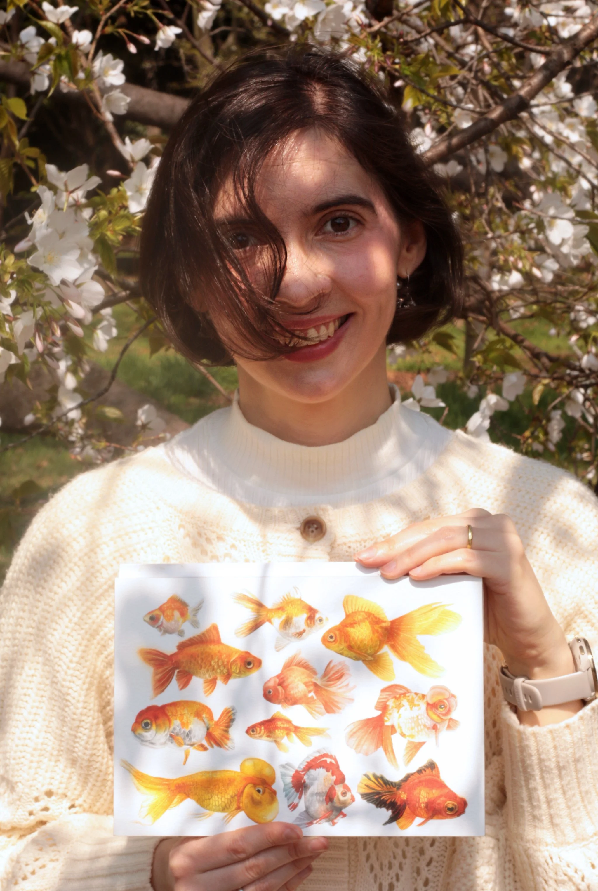 A woman with short dark hair smiling and holding a sheet of colorful fish illustrations in front of blooming tree branches with white flowers.