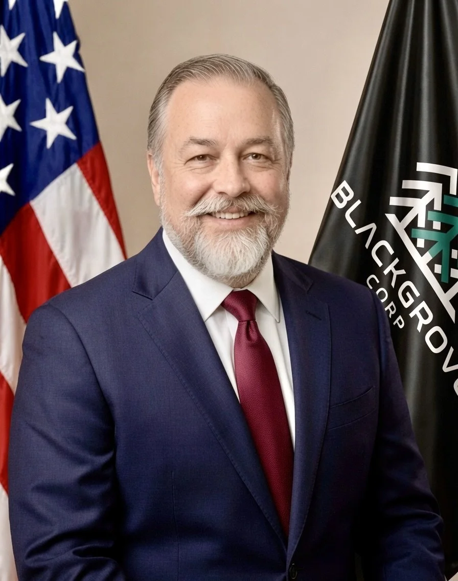 A man with a beard in a navy suit with a red tie, standing in front of an American flag and a BlackGrapevine Corp flag.