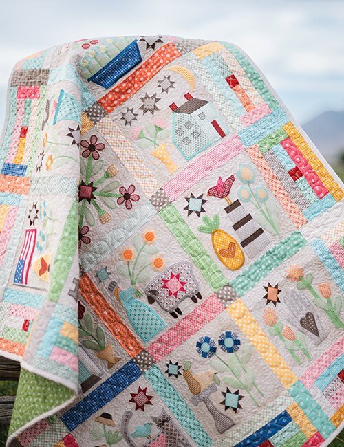 Quilt with various colorful patchwork patterns and appliqué designs, including flowers, animals, and houses, draped over a surface outdoors.
