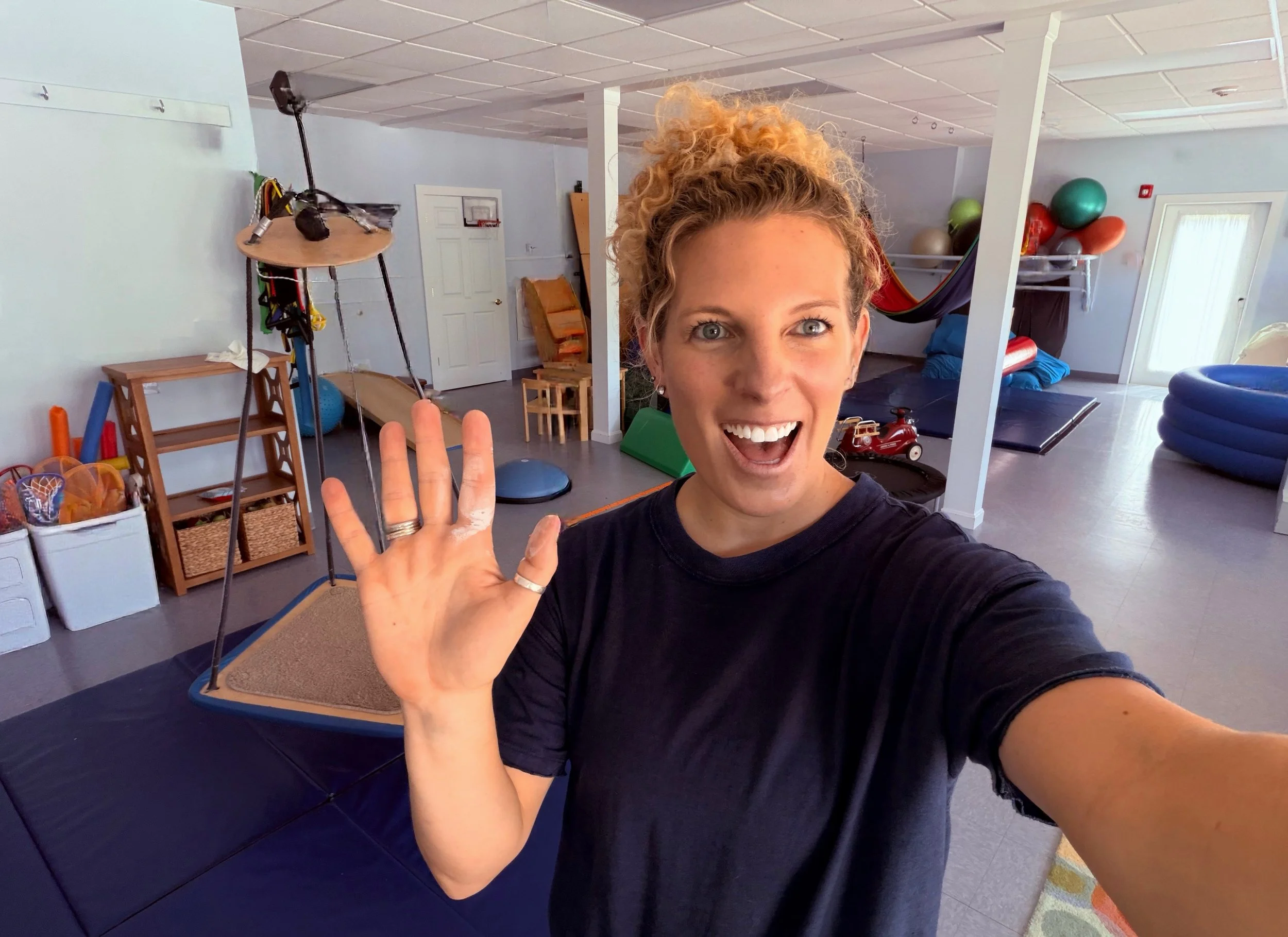 A woman with curly blonde hair smiling and waving in a room filled with workout equipment, including stability balls, yoga mats, and a basketball hoop on the wall.