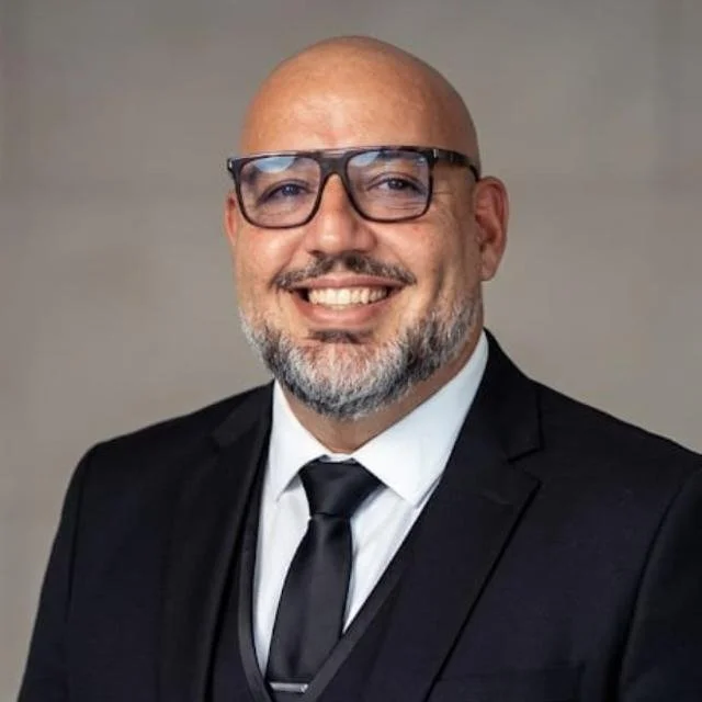 Homem de terno preto, camisa branca, sem cabelo, barba grisalha, usando óculos de armação escura, sorrindo.