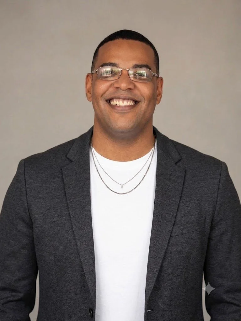 Homem sorridente usando óculos, blazer cinza e camiseta branca, de cabelos curtos, em um fundo neutro.