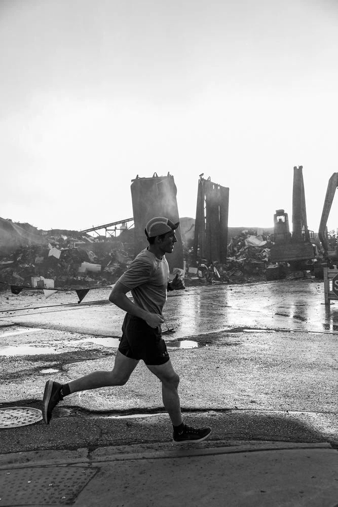 A man wearing a hat, t-shirt, and shorts jogging past a construction or demolition site with debris and ruined structures in the background.