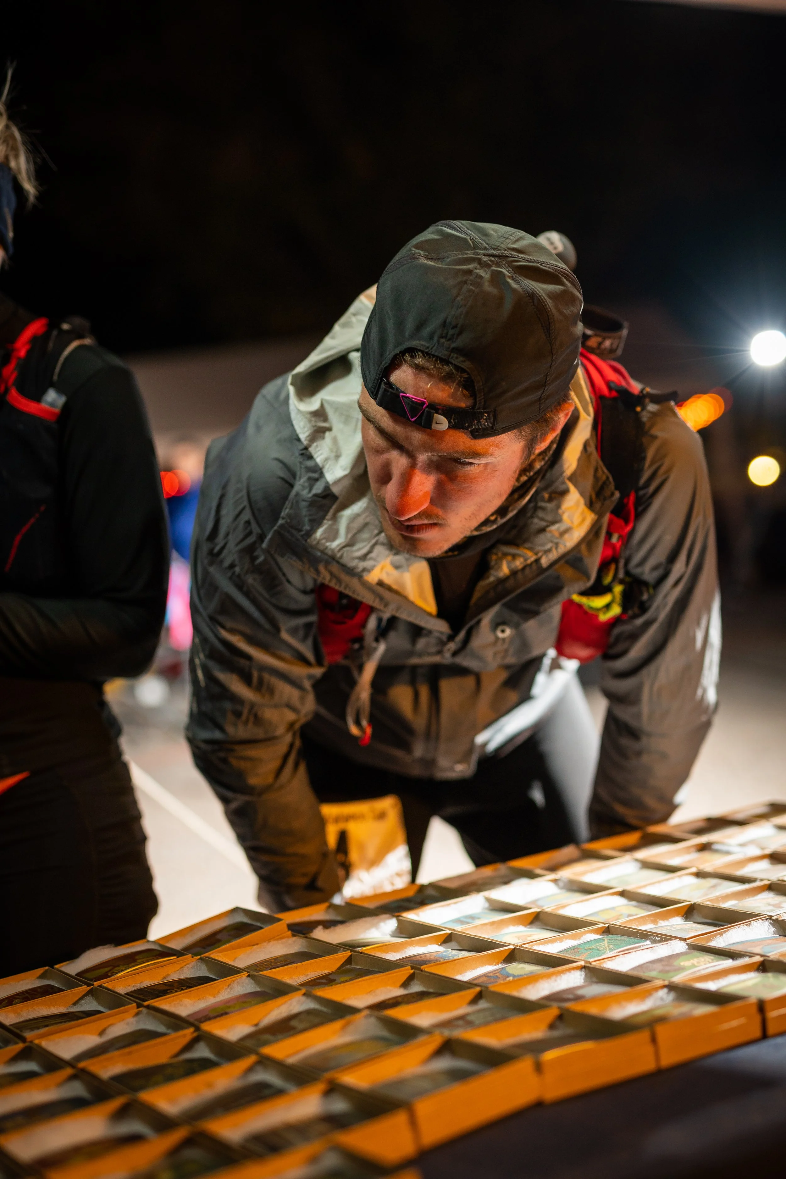 A man wearing a cap and outdoor gear examining a display of colorful items on tables at night.