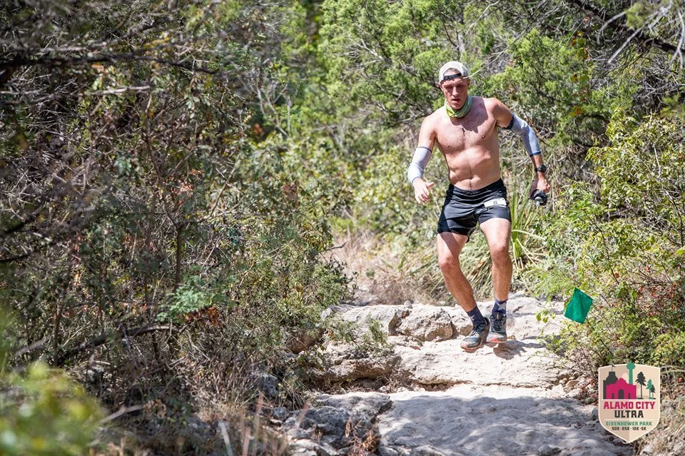 A shirtless man wearing black shorts, sunglasses, and a headband running on a rocky trail through a green, bushy outdoor area. There is a green flag marker on the side of the trail, indicating it is part of an organized event, the Alamo City Ultra.