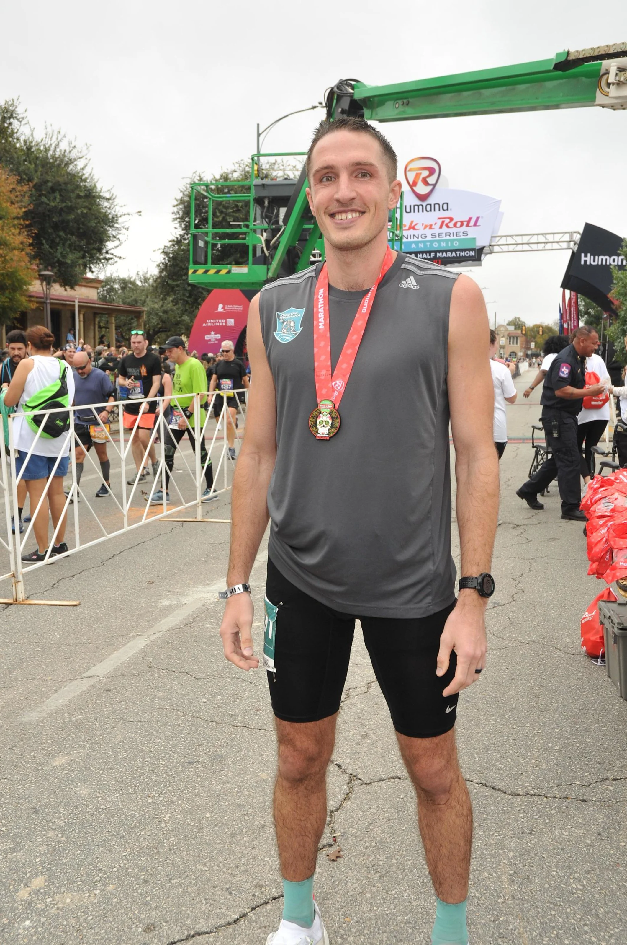 A smiling man in running attire with a medal around his neck stands at a marathon finish line surrounded by other runners and event staff.
