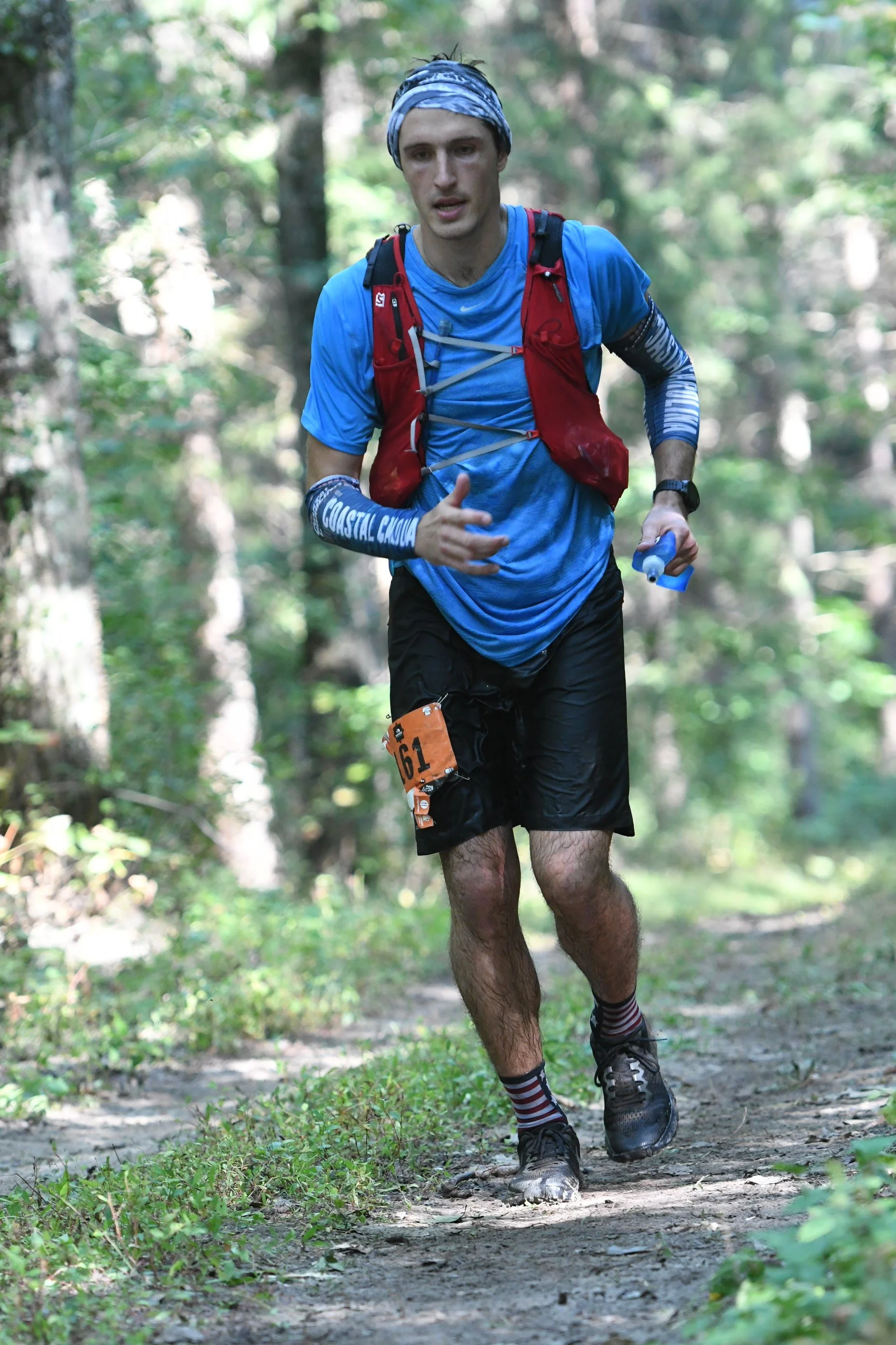 A man running on a trail in a forest with trees and greenery, wearing a blue shirt, black shorts, a red backpack, and striped socks.