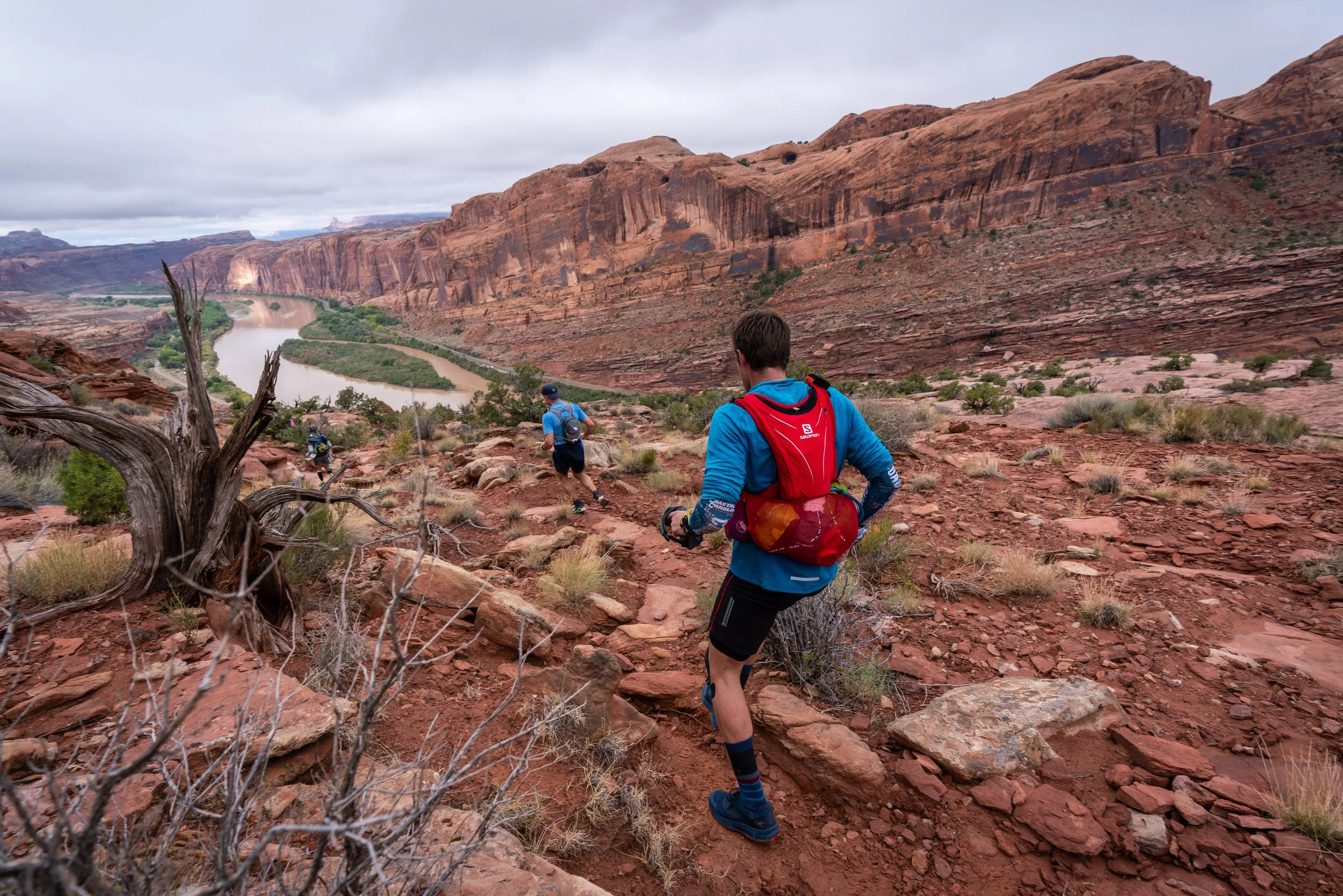 Trail runners on a rocky trail overlooking a winding river and red rock canyon under cloudy sky in a desert landscape.