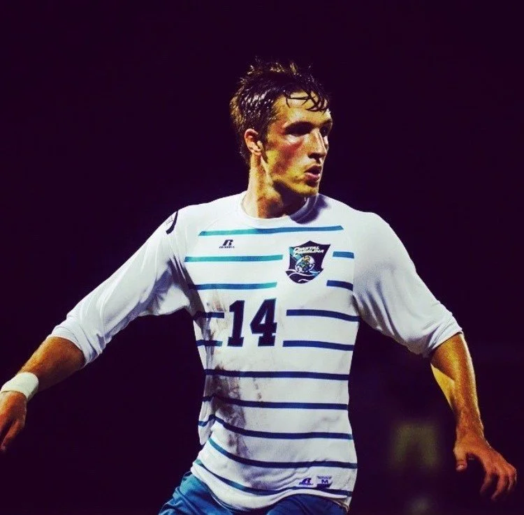 Male soccer player in white and blue striped jersey with number 14 and a team crest on the chest, standing on the field at night.
