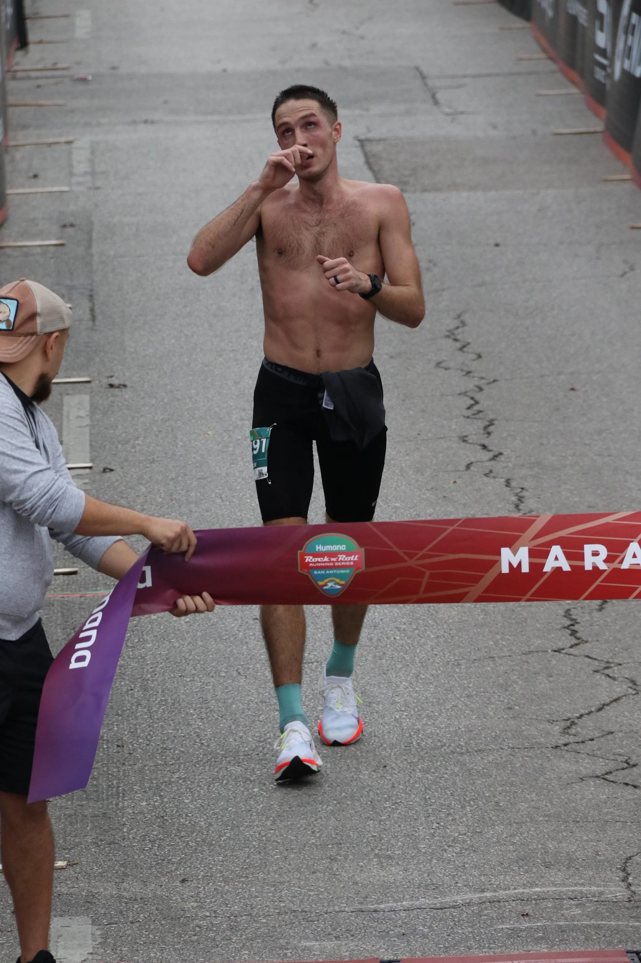 A shirtless man with short dark hair and a race bib crosses the finish line of a marathon, wiping his nose with his hand.