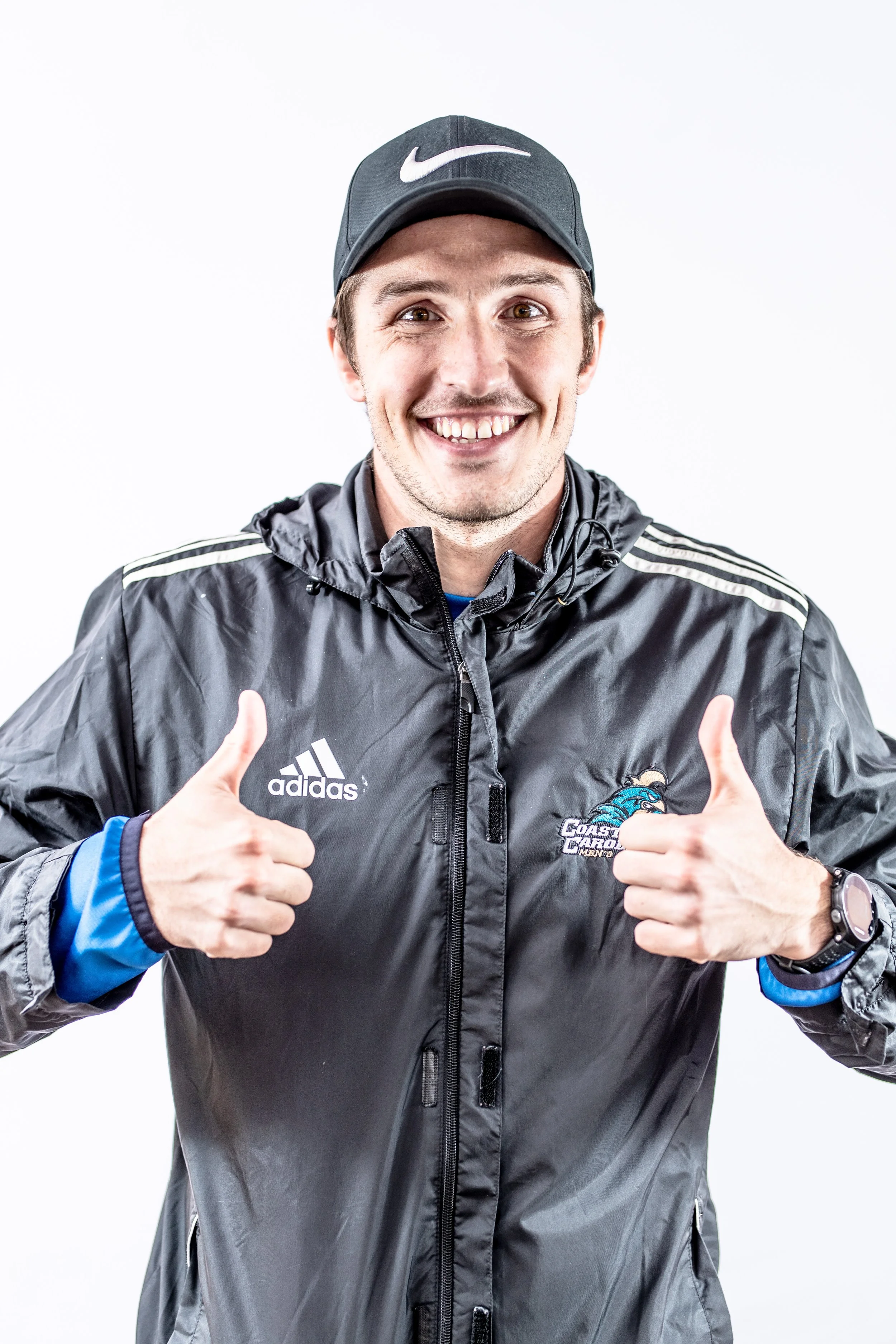 A smiling man wearing a black Nike cap, black Adidas and Coast Guard all-star jacket, showing thumbs up.