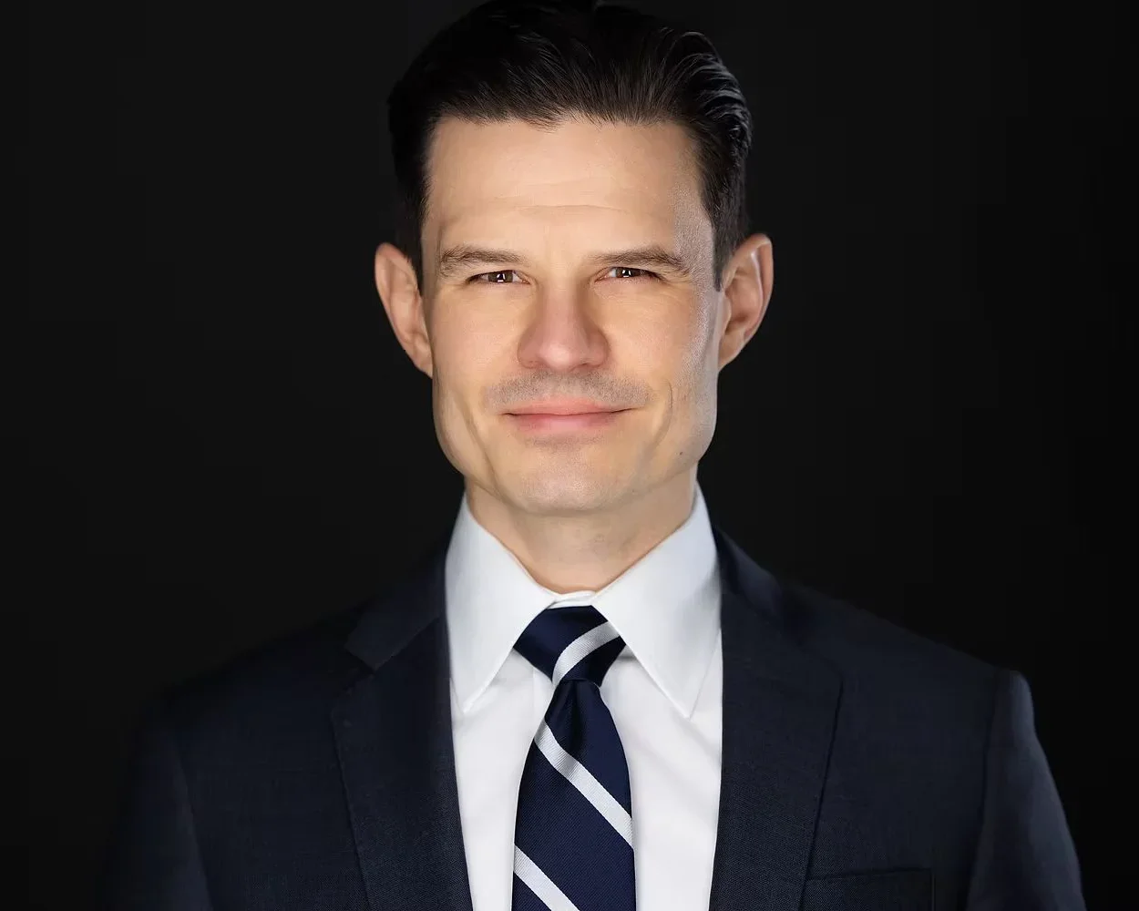 Headshot of a smiling Michael Willes in a black suit, white shirt, and striped tie against a black background.