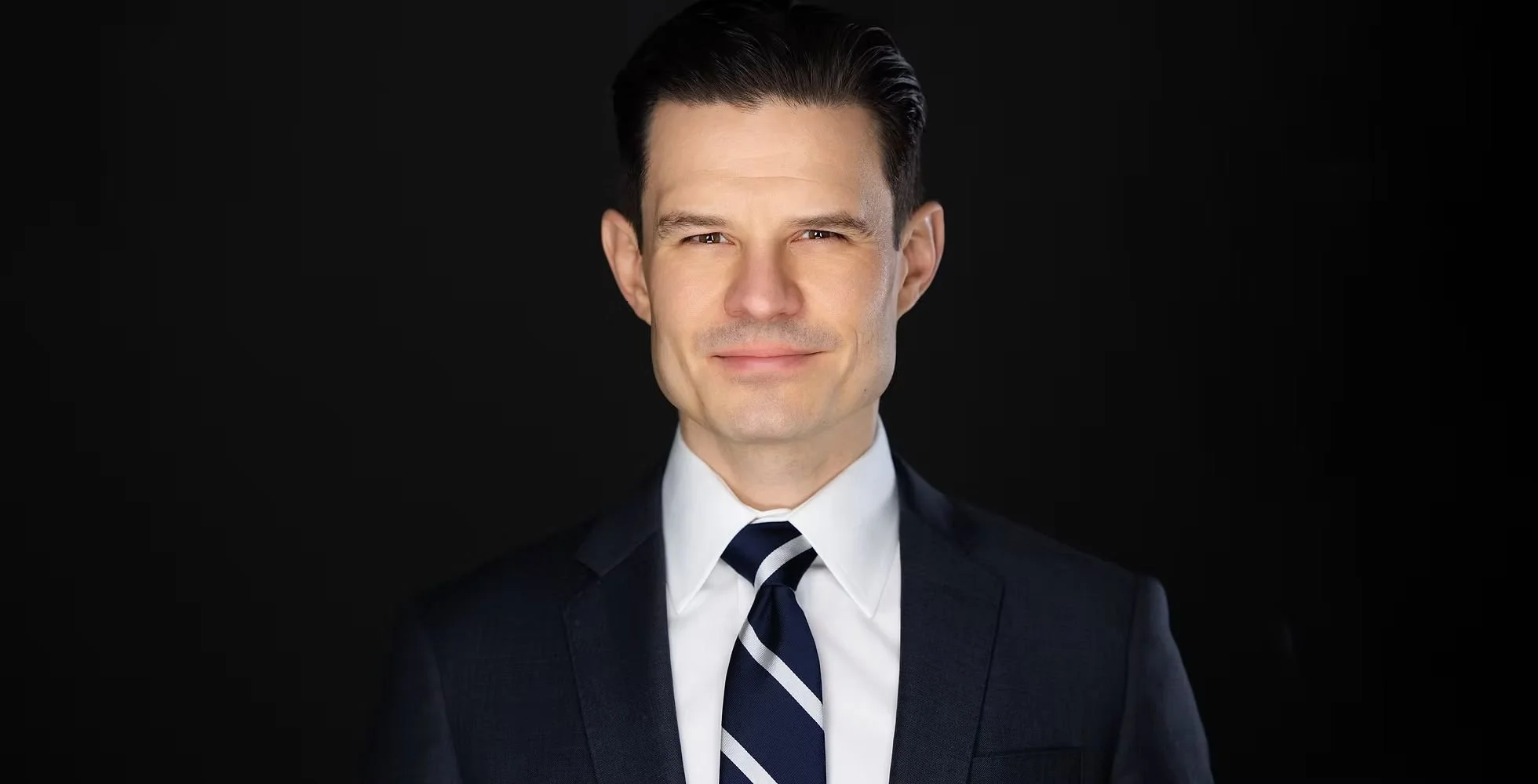 Portrait of Michael Willes with dark hair, wearing a dark suit, white shirt, and a striped tie, against a black background.