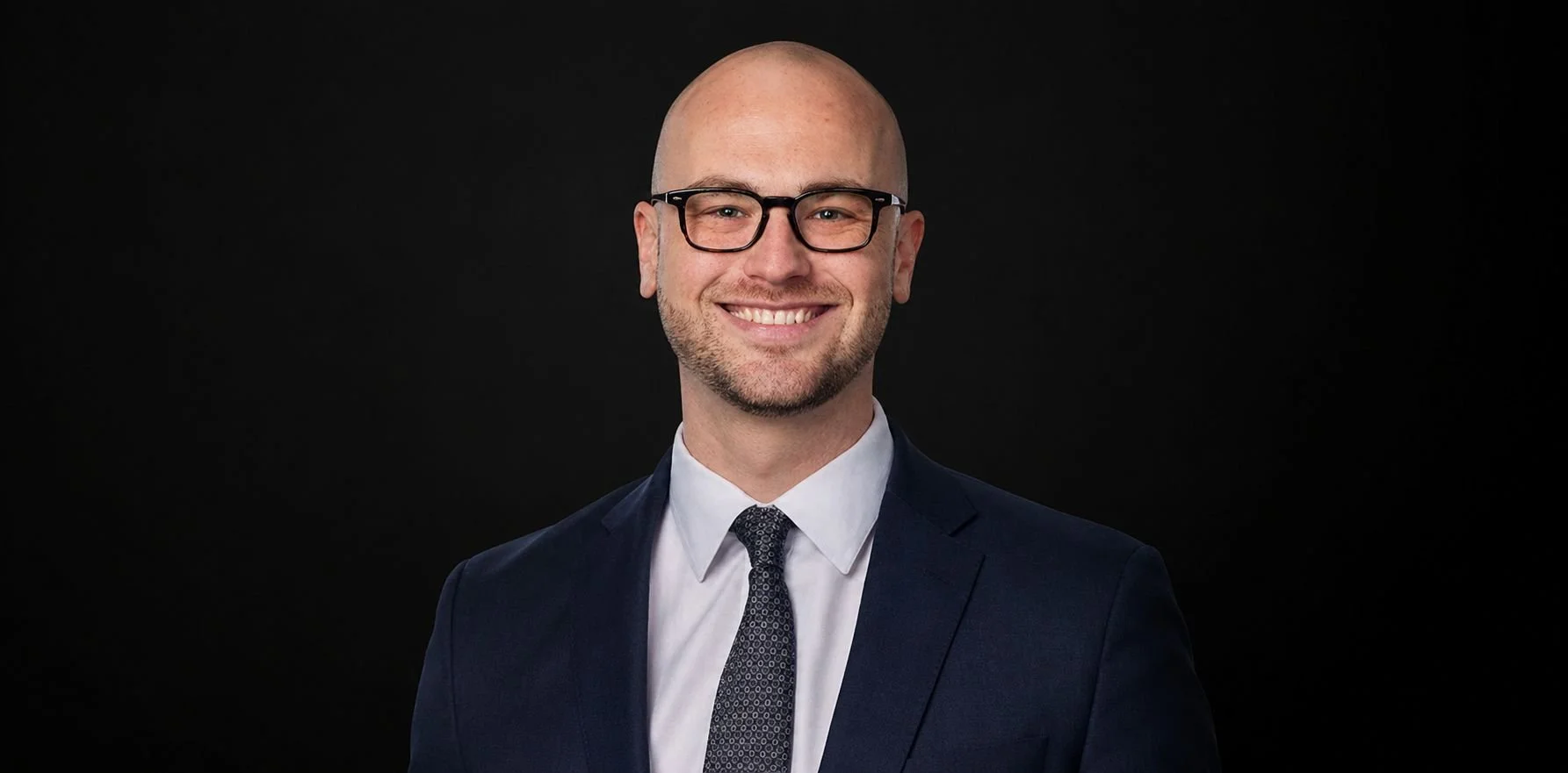 Portrait of Joshua Masser with glasses, wearing a navy blue suit, white shirt, and a patterned tie, set against a black background.