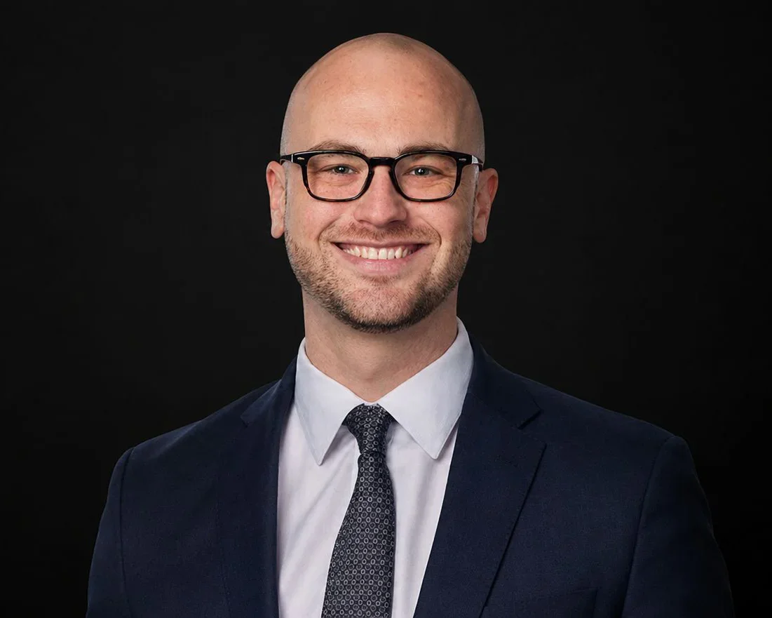A smiling Joshua Masser with glasses, wearing a dark suit, white shirt, and patterned tie, against a black background.