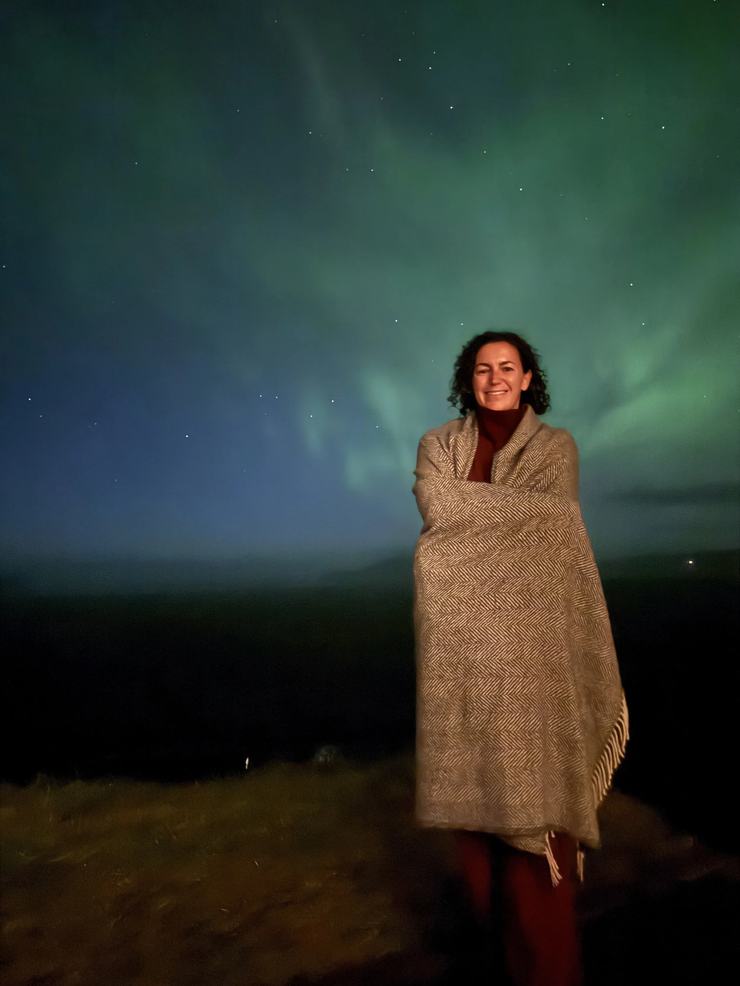 A woman with dark curly hair smiling and crossing her arms, standing outdoors at night, with the Northern Lights in the sky behind her.