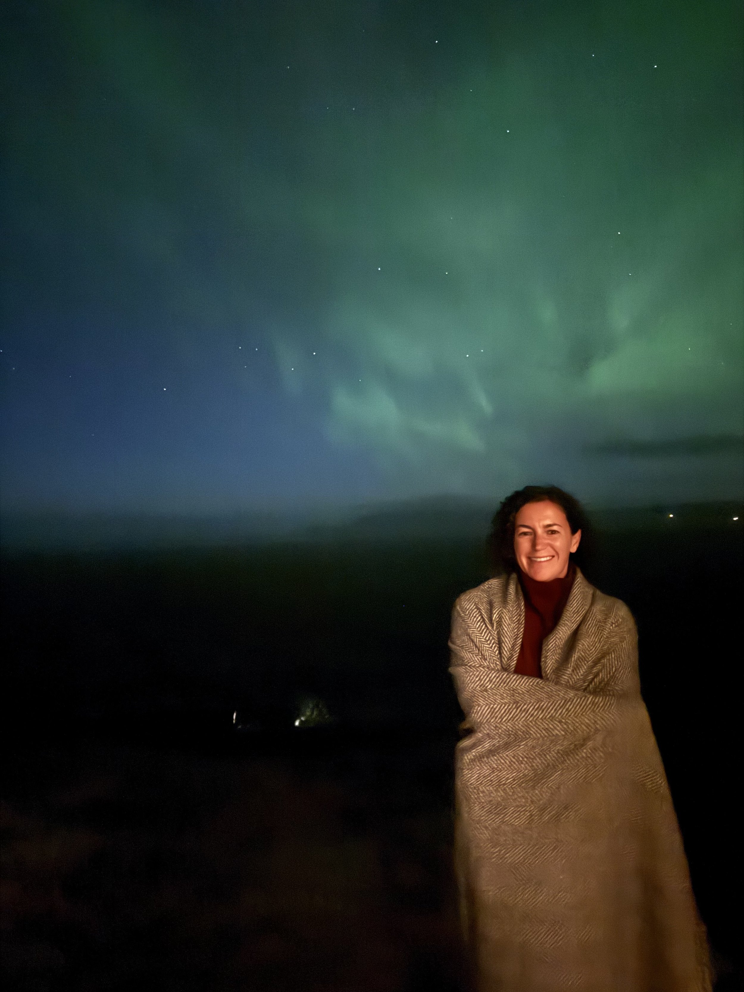 A woman standing outdoors at night with a bright smile, wearing a patterned coat, with the northern lights visible in the sky above.