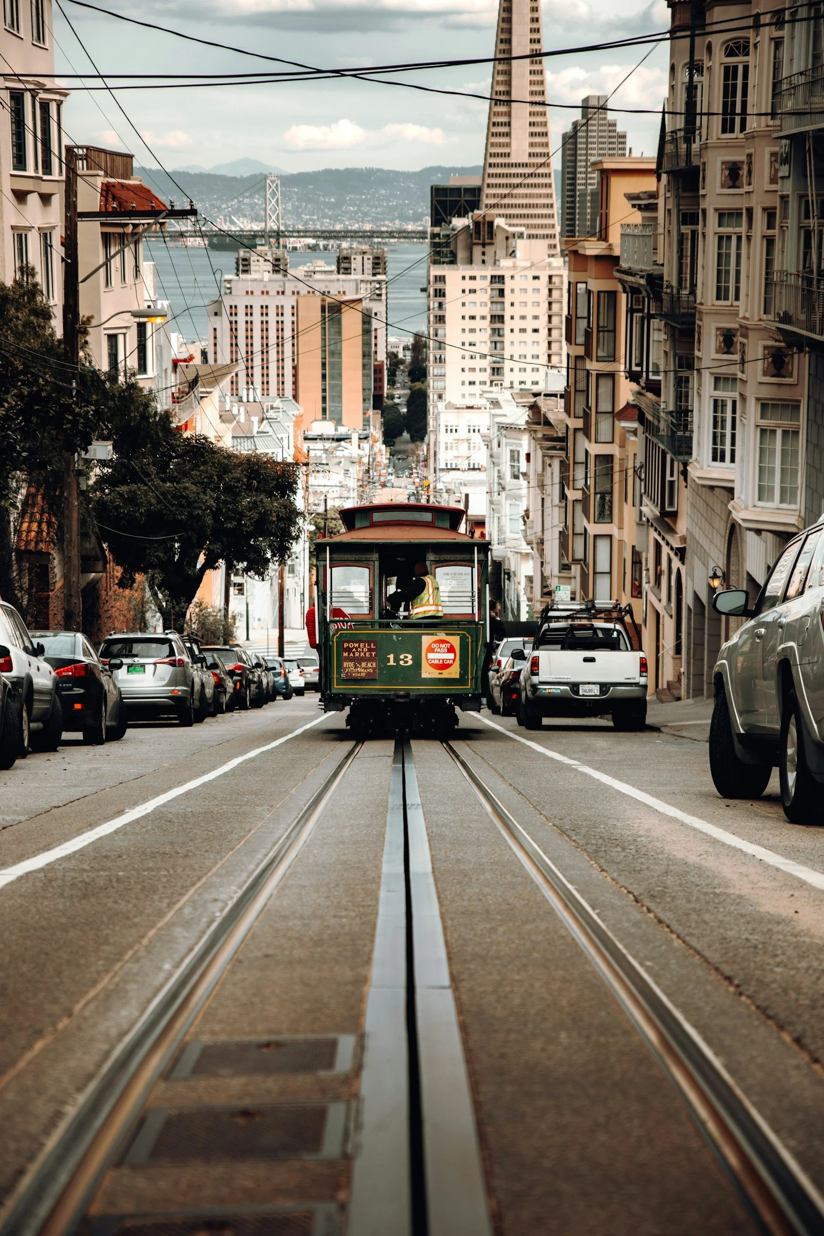 View of a city street with cable car in the center, surrounded by parked cars on both sides, with tall buildings in the background, including the Transamerica Pyramid and the Bay Bridge in San Francisco. The sky is cloudy.