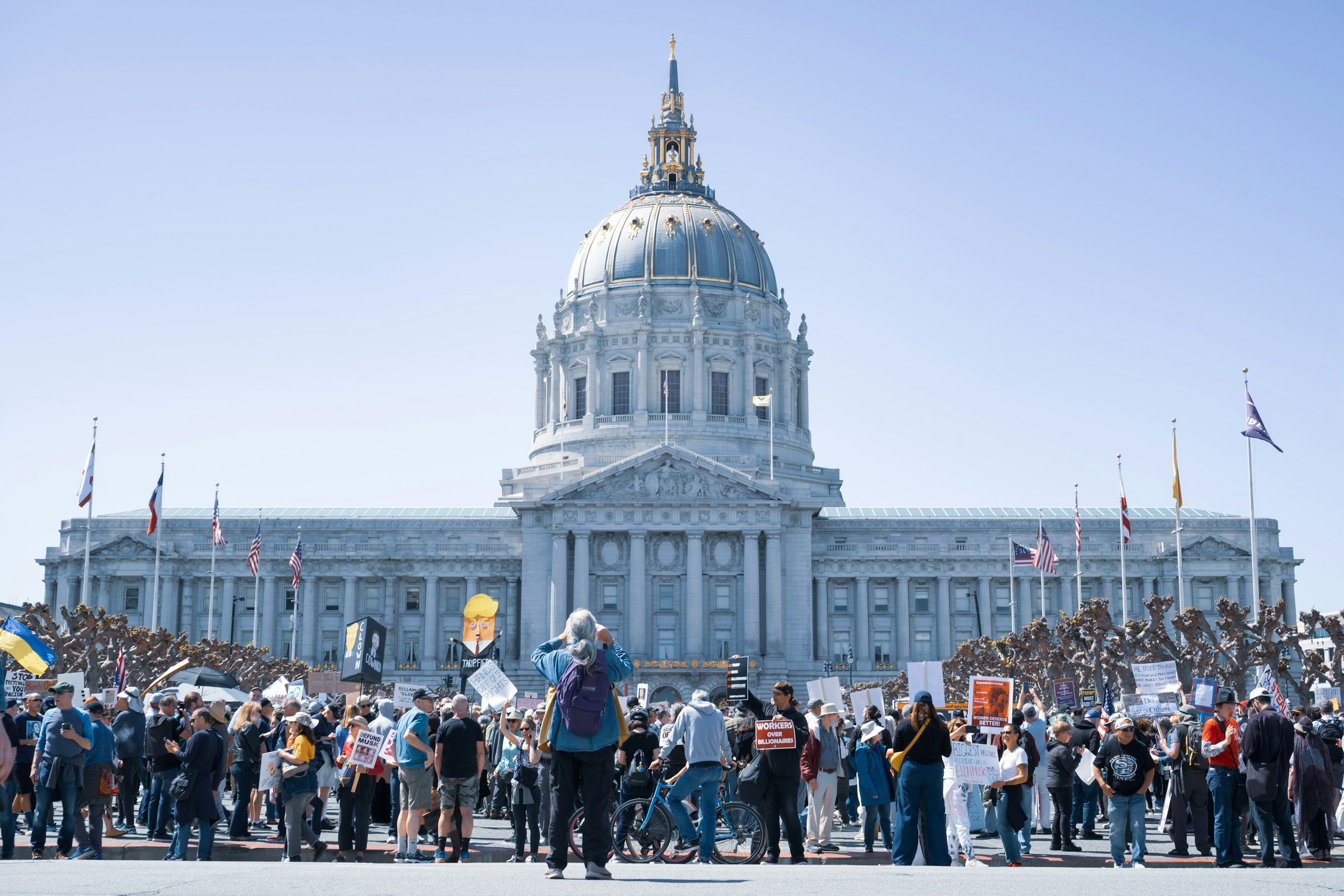 A large crowd of protesters gathered in front of a government building with a dome, holding signs and banners. The building has a classical architecture style with flags flying in front.