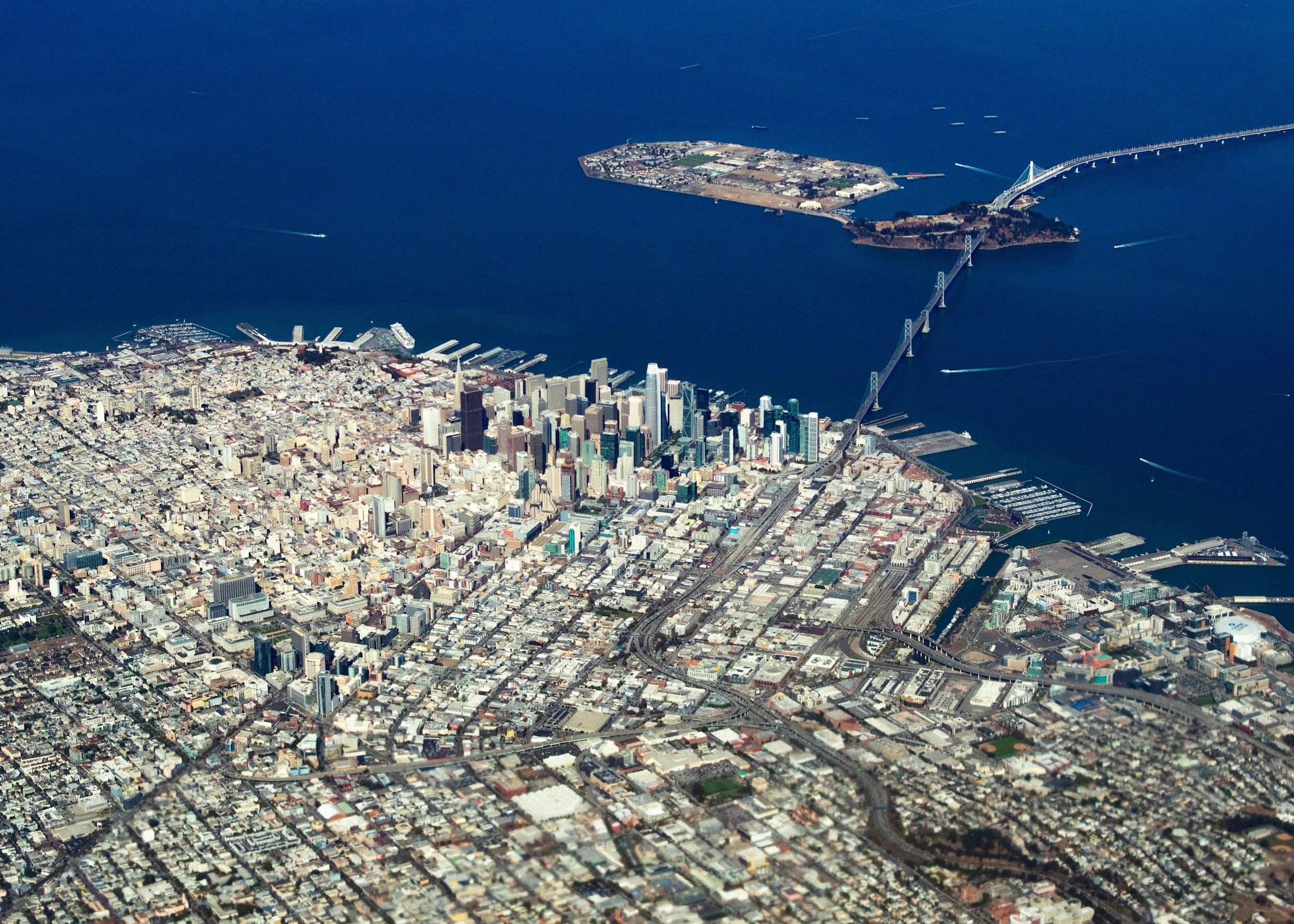 An aerial view of a city with a large bay, a bridge crossing over the water, and a small island in the bay. The city is densely packed with buildings, roads, and some green spaces.