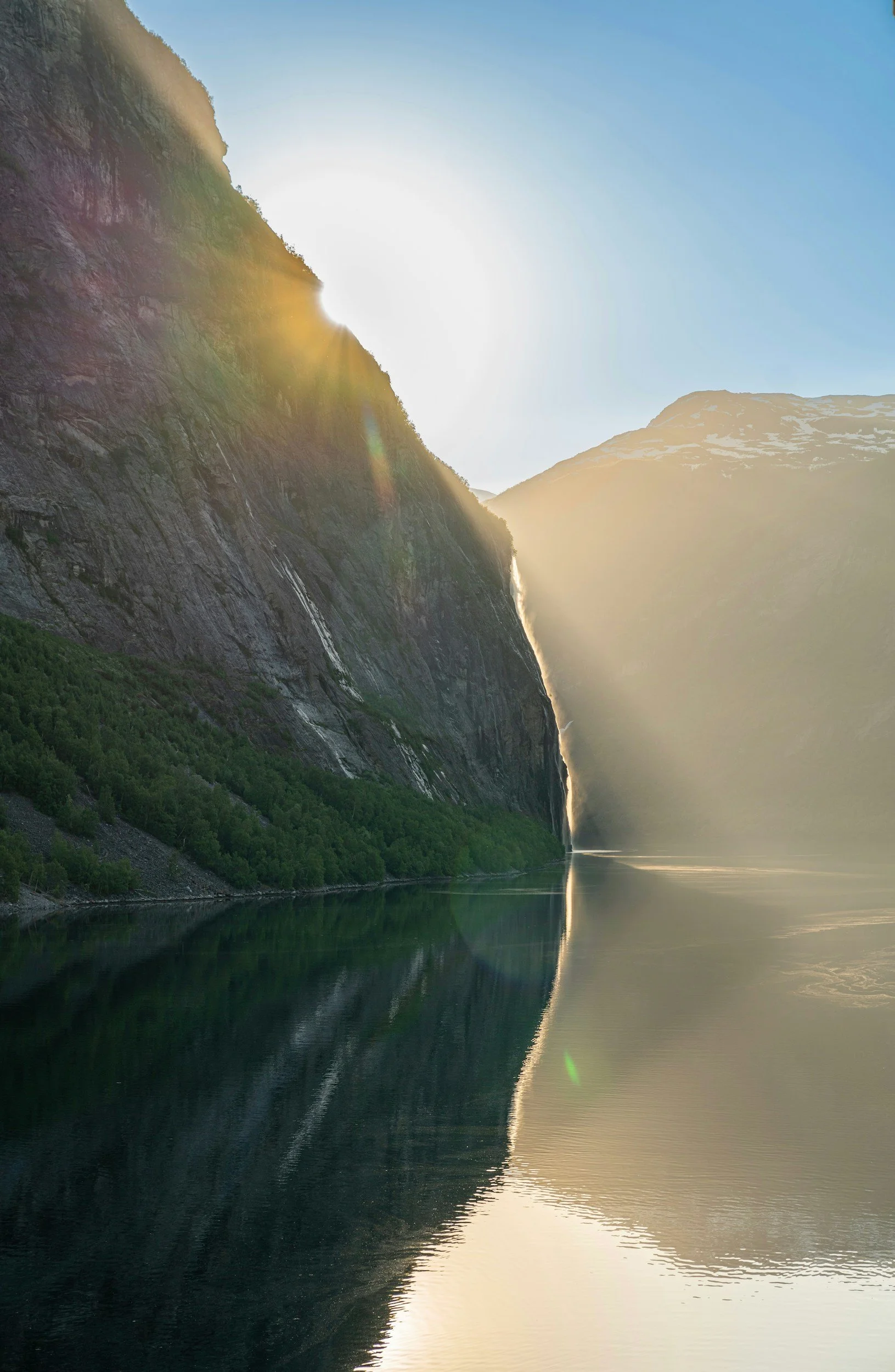 Sunlight shines over a large, rocky mountain with a tall waterfall cascading down its side. The mountain is reflected in a calm body of water at its base, with a clear blue sky above.