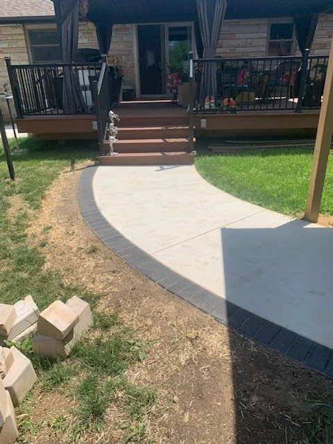 Backyard with a wooden deck, stairs, and a concrete patio with stone edging, grass, and some bricks.