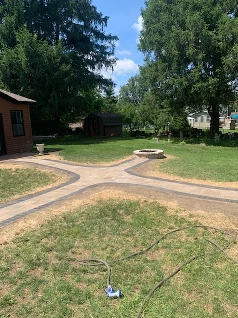 Backyard with grass, trees, and a brick walkway forming a cross shape, a small fire pit, and a garden hose on the ground.