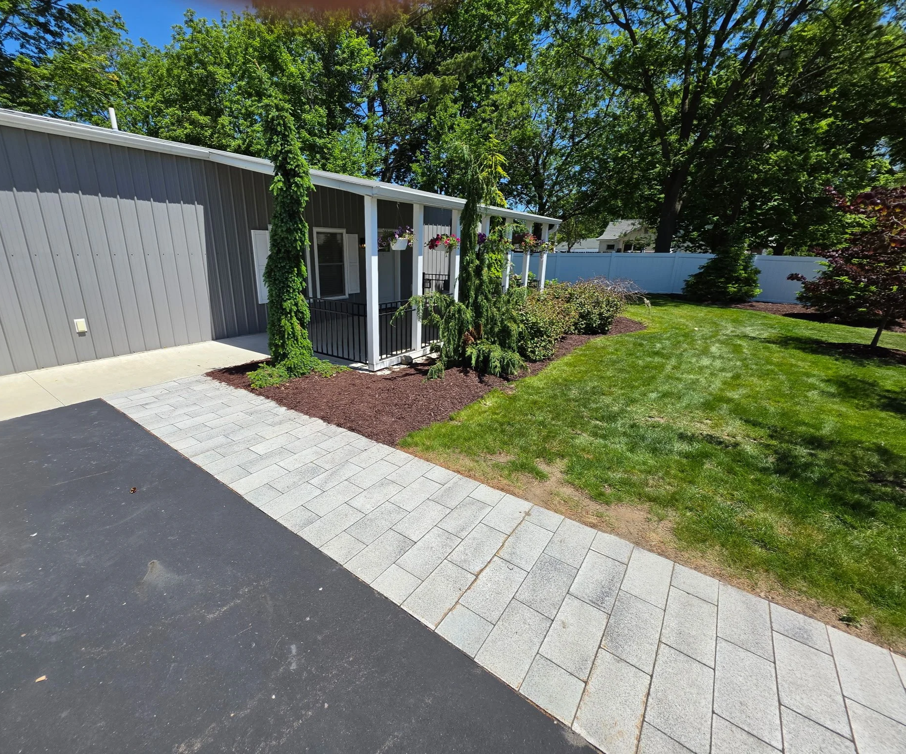 A backyard scene with a gray metal building, a small porch with hanging flower baskets, a neatly mowed green lawn, and trees along a white privacy fence on a sunny day with a blue sky.