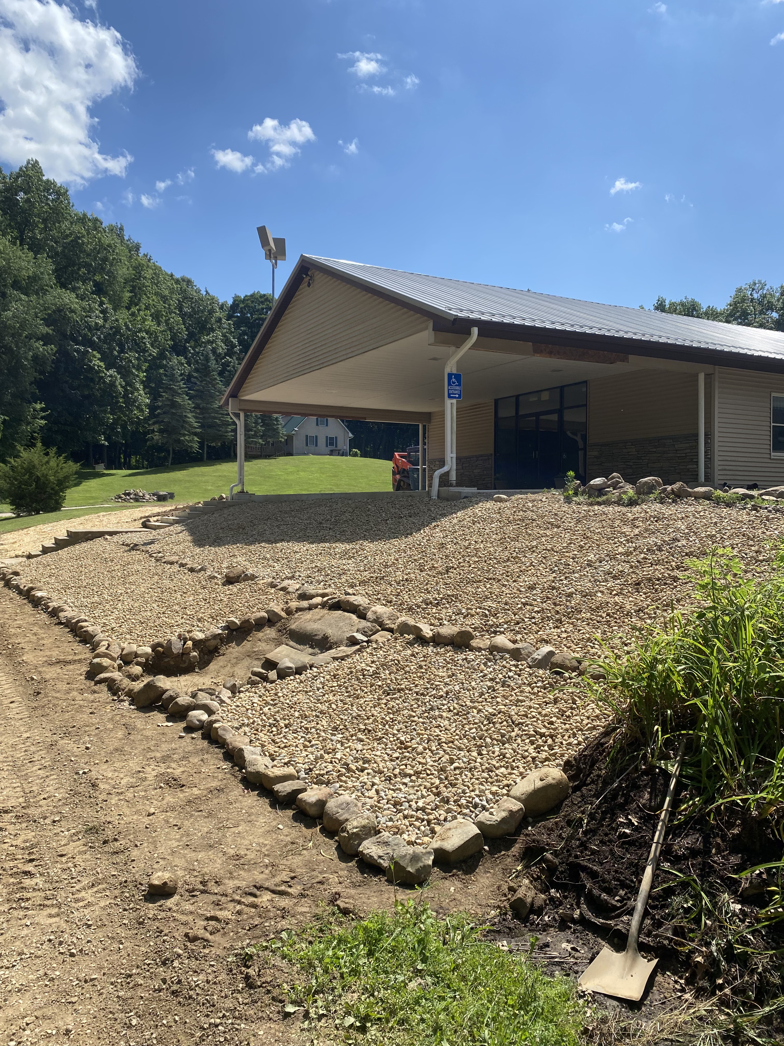A house with a sloped roof and a gravel parking area in front, with a small landscaped patch edged with rocks, a shovel leaning against a plant, and a sign indicating accessible parking spaces, during daytime with a bright blue sky and some clouds.