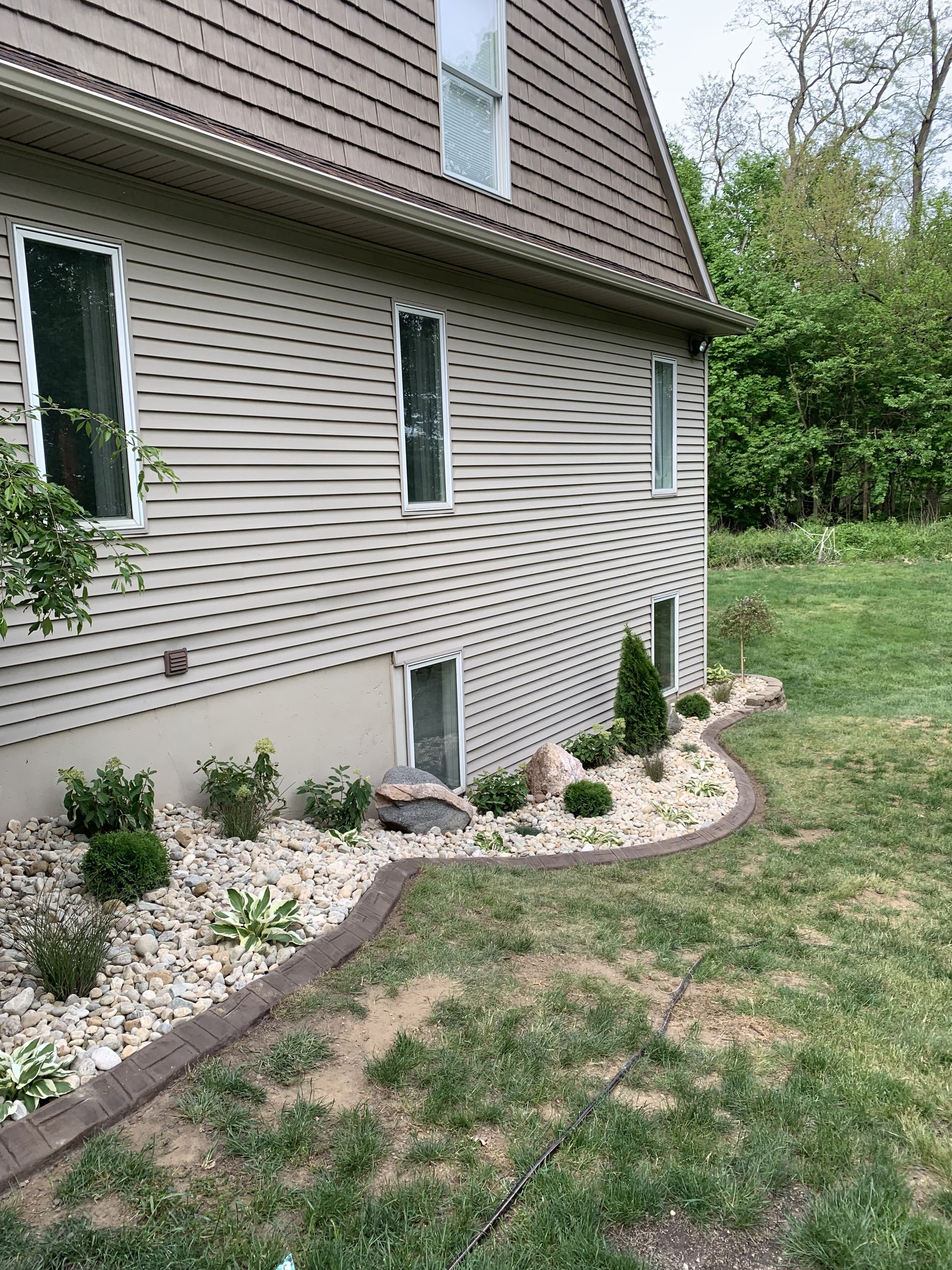 The side of a house with beige vinyl siding and several windows, some with blinds. There is a landscaped area with rocks, small bushes, and plants bordered by a curved brick edging. The grass nearby is patchy with some bare spots.