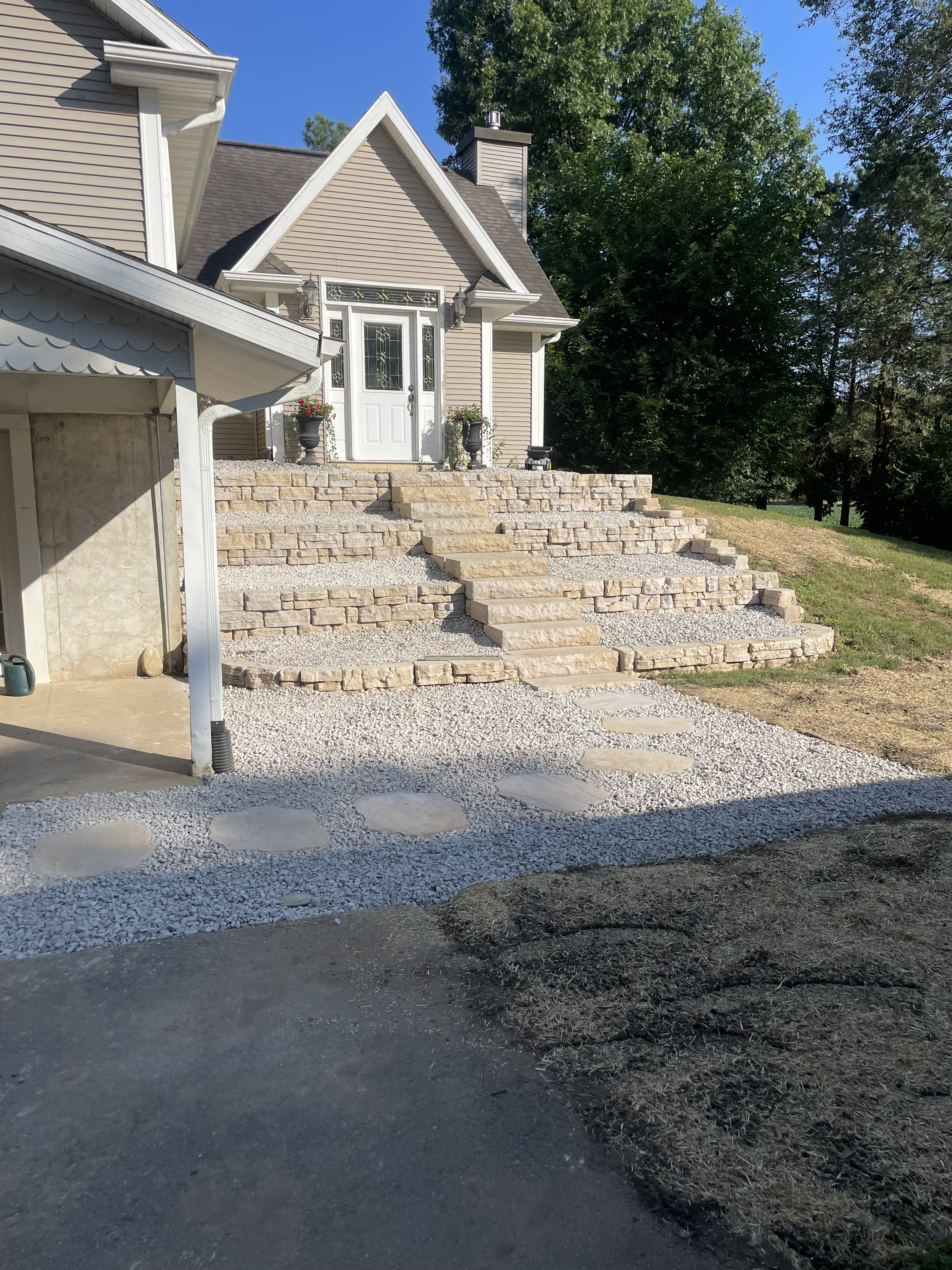 Front steps of a house with stone stairs and gravel pathway, flanked by potted plants, with a house and green trees in the background.