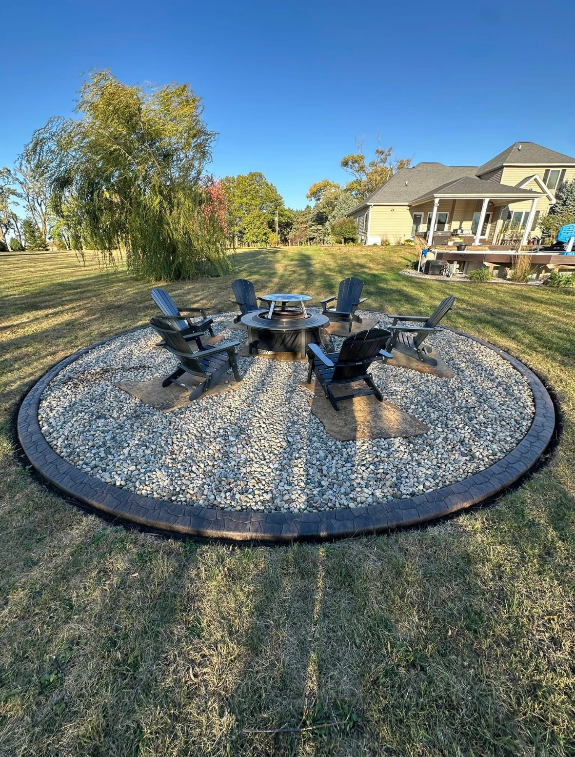 A round outdoor fire pit seating area with six black Adirondack chairs on a gravel and stone circle in a backyard.