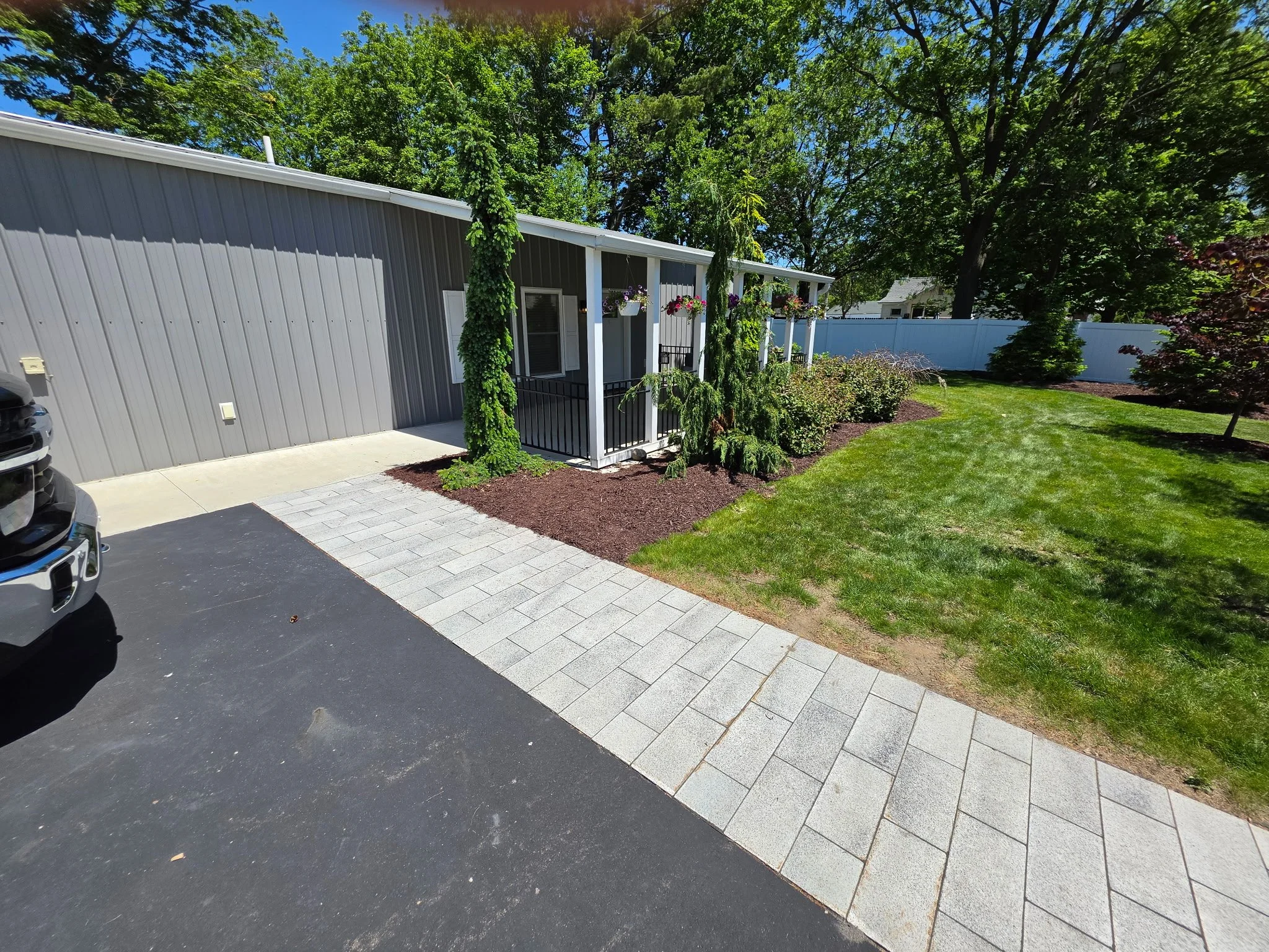 A backyard with a gray house, lush green trees, a well-maintained lawn, and hanging flower baskets. There is also a black and gray vehicle partially visible in the driveway.