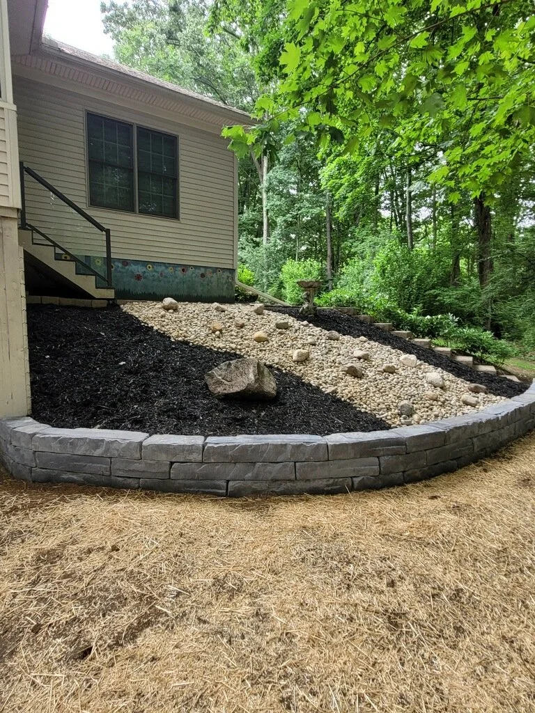 A backyard landscape with a house on the left, a tiered stone retaining wall, and a garden bed with mulch, rocks, and a bird bath surrounded by trees.