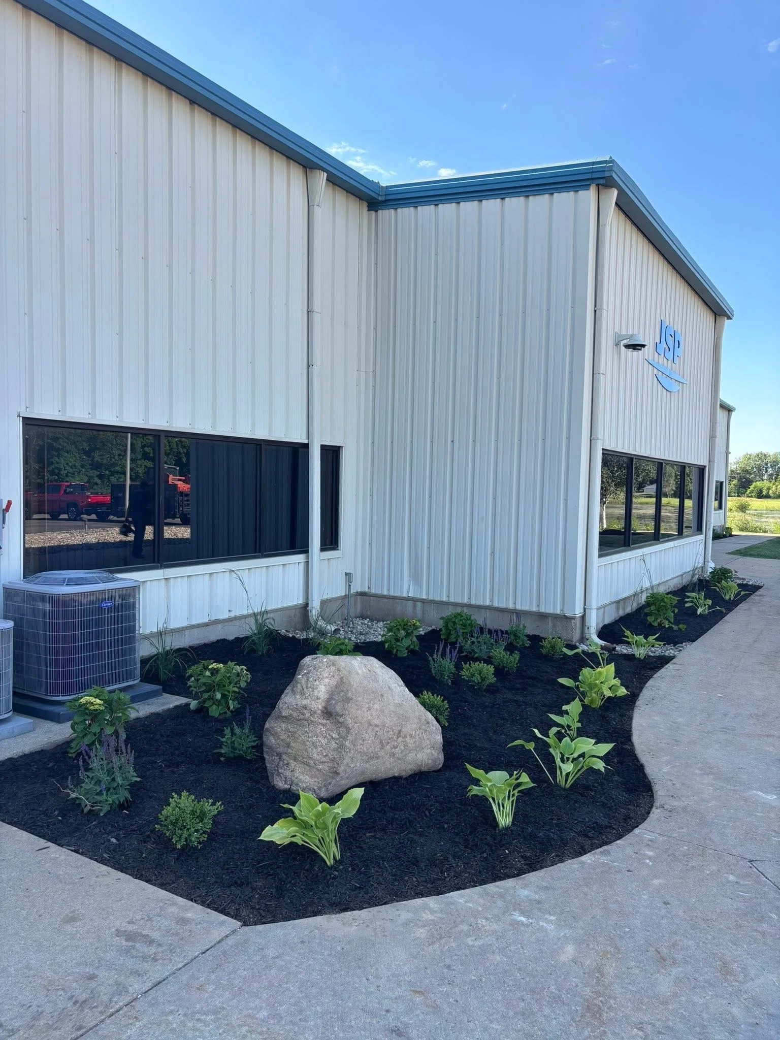 Exterior of a commercial building with white metal siding, black framed windows, a small landscaped garden bed with green plants and a large rock, and an air conditioning unit outside