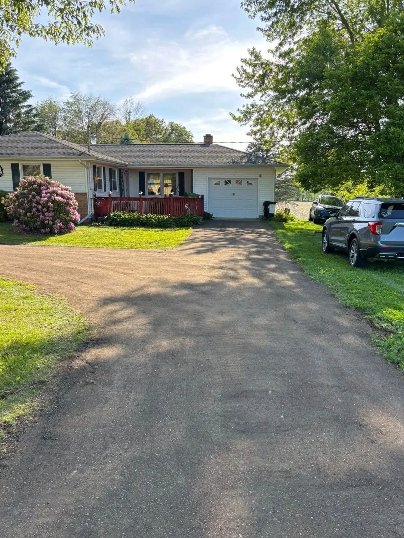 A house with a driveway, parked cars, green lawn, trees, and a blue sky with some clouds.