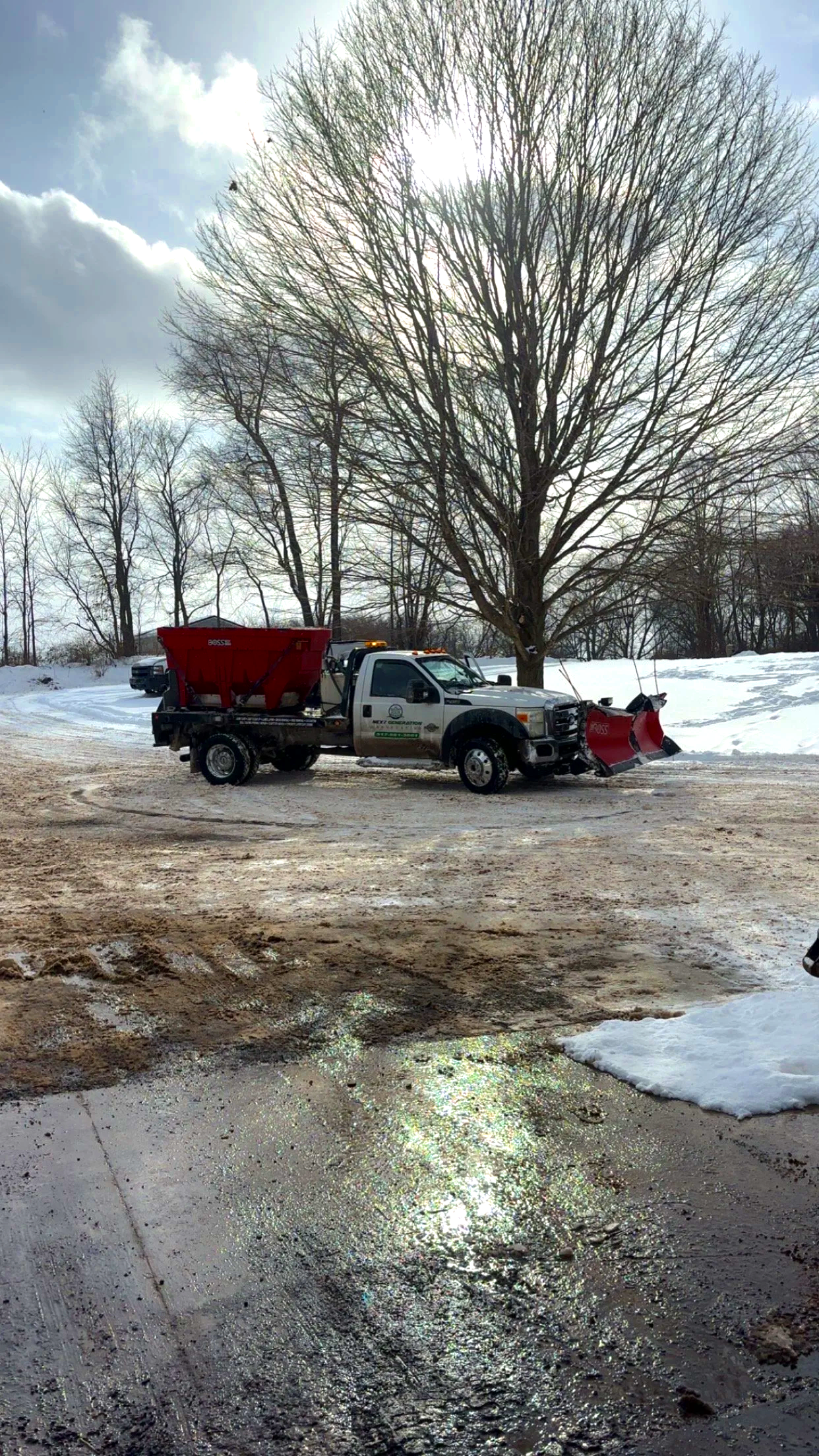A snow-covered parking lot with a utility truck equipped for snow removal, parked in front of a large leafless tree on a cloudy winter day.