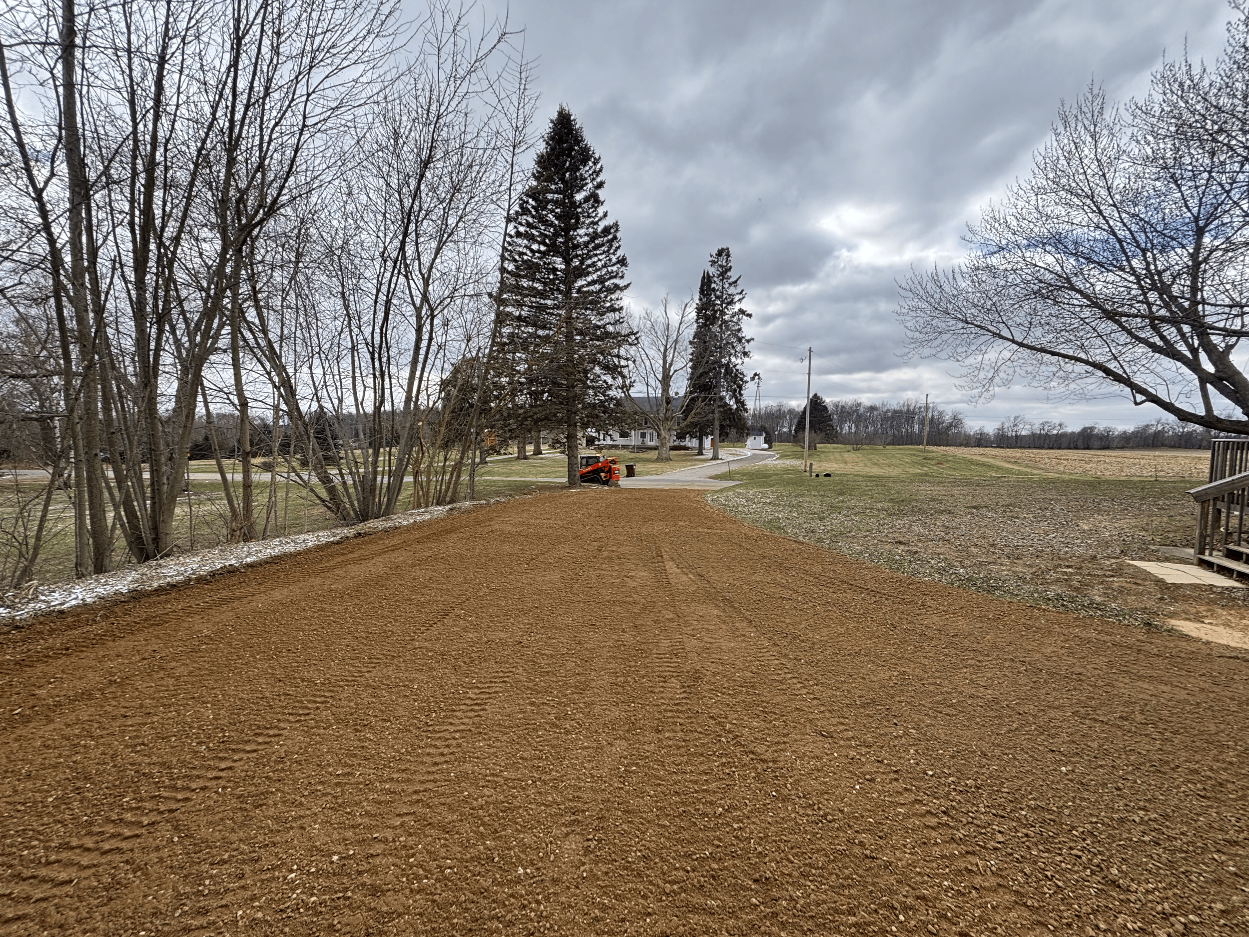 A rural scene with bare trees, a cloudy sky, a snow plow truck on the street, a house, a pasture, and power lines.