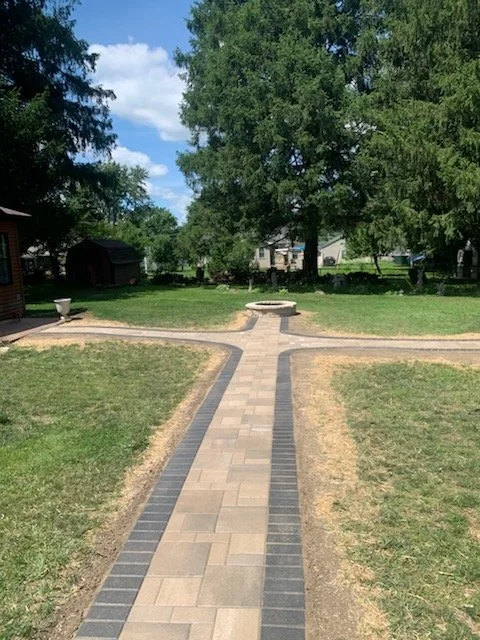 A paved walkway with a cross shape that leads to a fire pit, surrounded by grass and trees in a backyard.