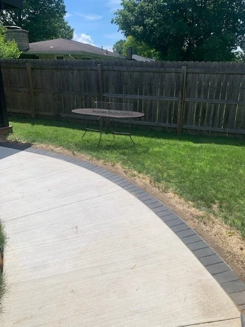 Backyard with a curved concrete patio, a wooden fence, a small table with folding legs, and trees in the background under a partly cloudy sky.