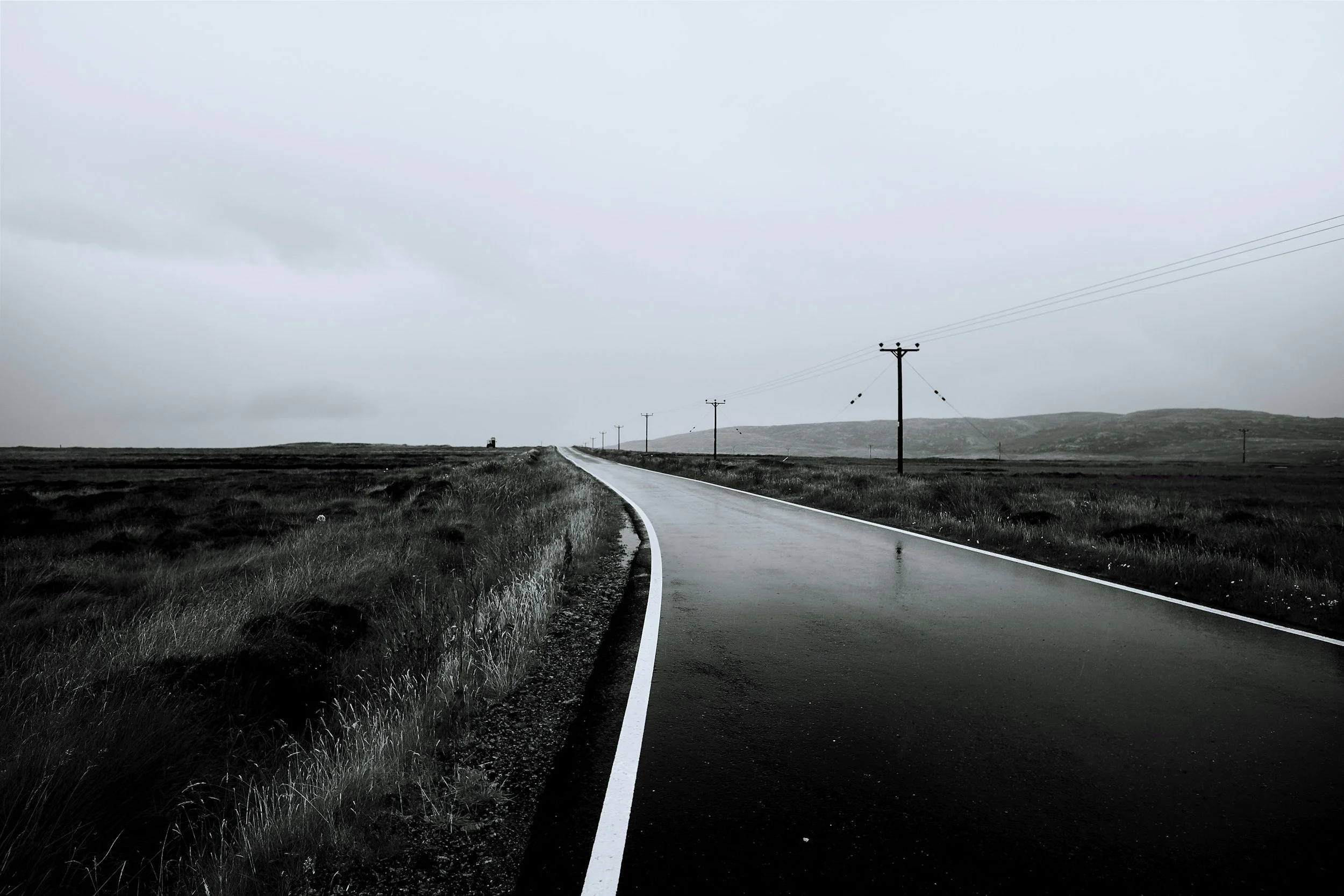 A long, winding road through flat grassland with hills in the distance, and a series of power lines running alongside it under a cloudy sky.
