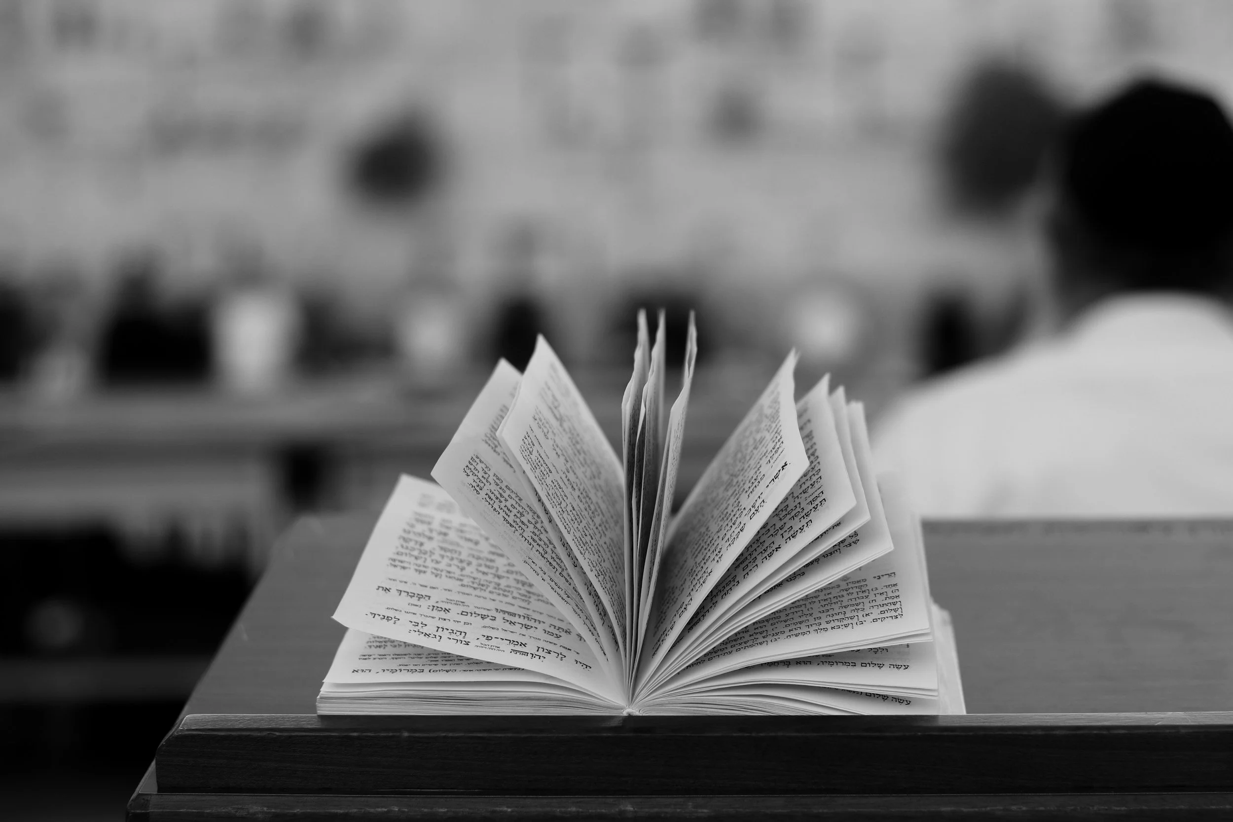 Open book with text in Hebrew, placed on a wooden table with a blurred background.