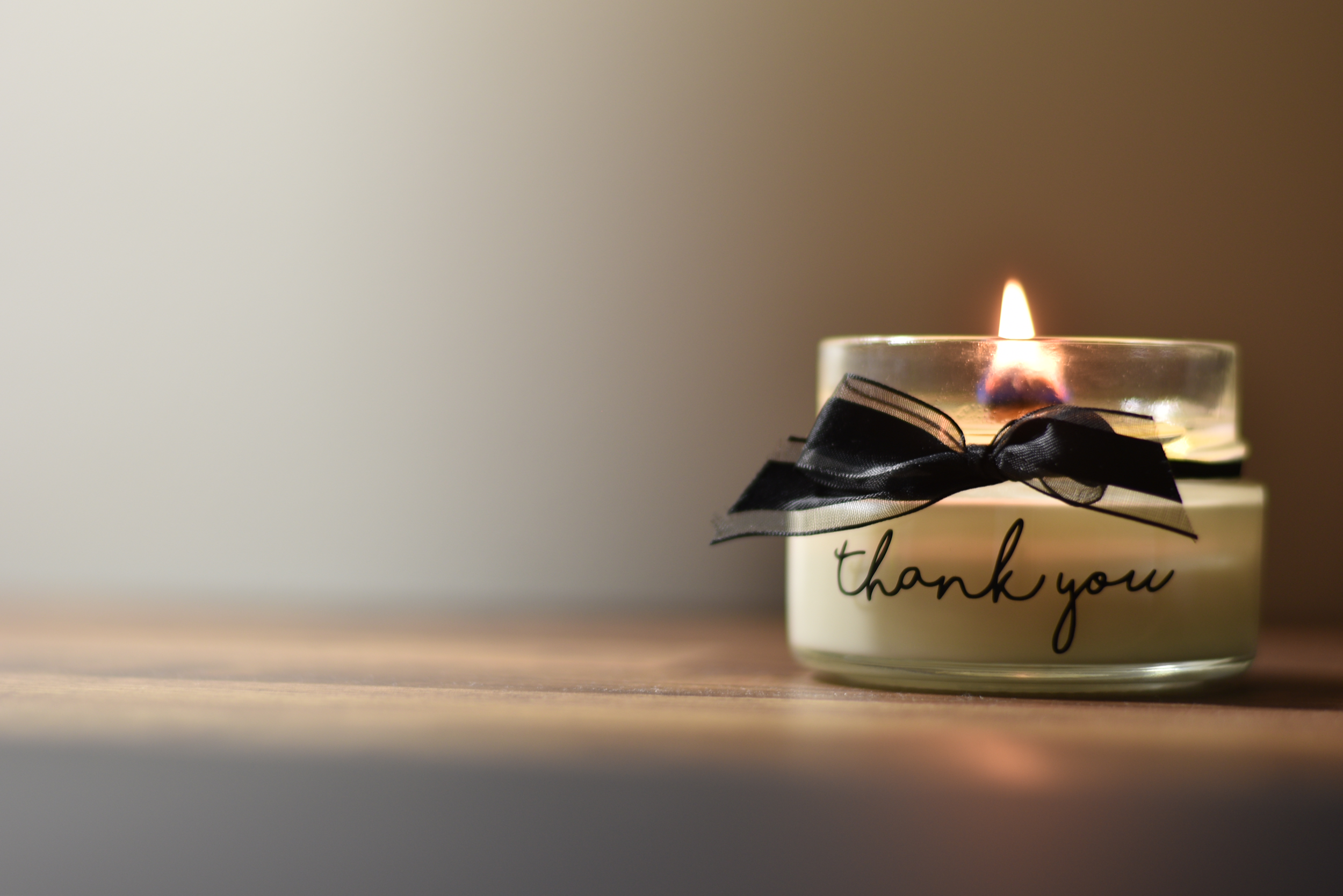 A glass candle holder with a lit candle inside, decorated with a black ribbon bow and a thank you message.