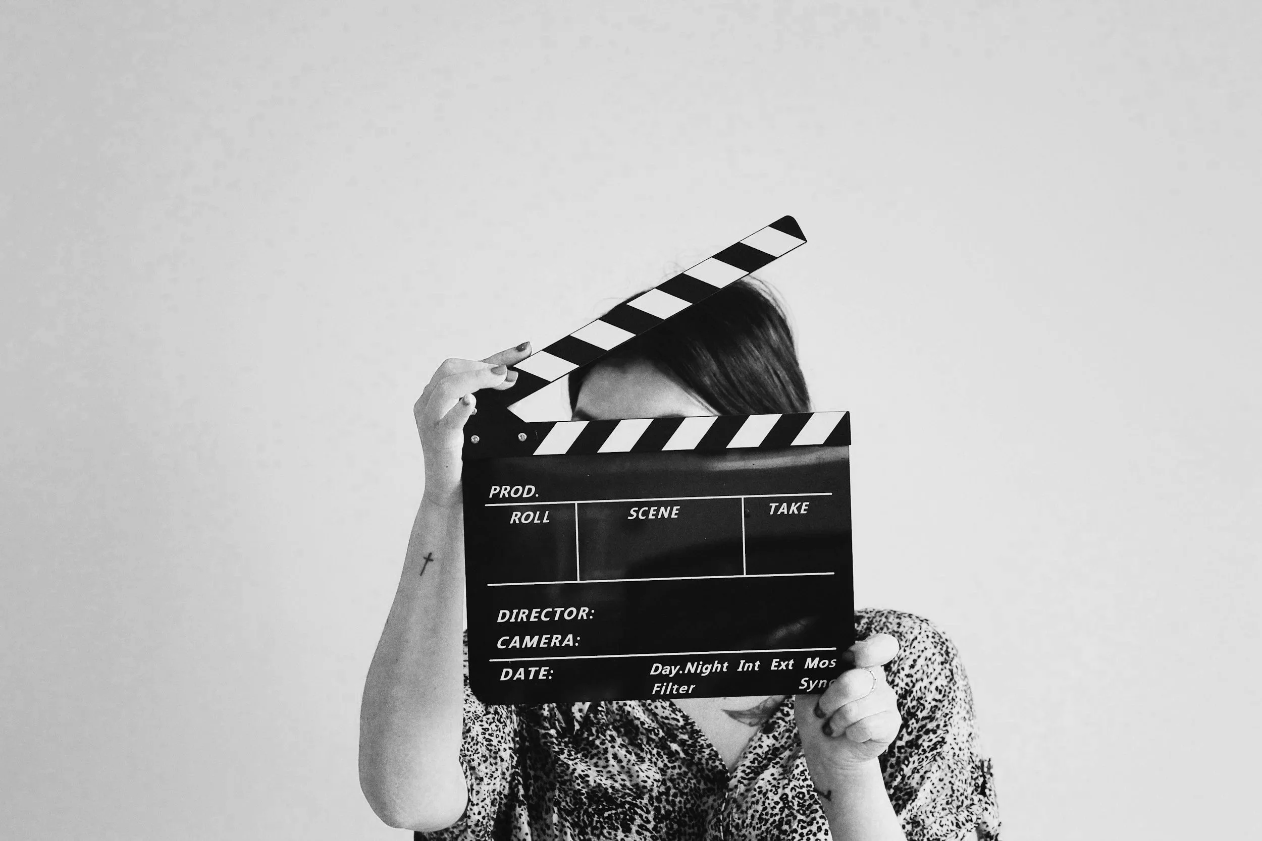 A person holding a film slate in front of their face. The person has dark hair and is wearing a leopard print top. The background is plain and light-colored.