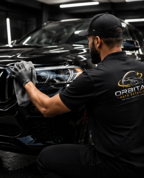A man wearing a black cap, black gloves, and a black t-shirt with an auto detailing logo is polishing a black car in an indoor auto detailing shop.