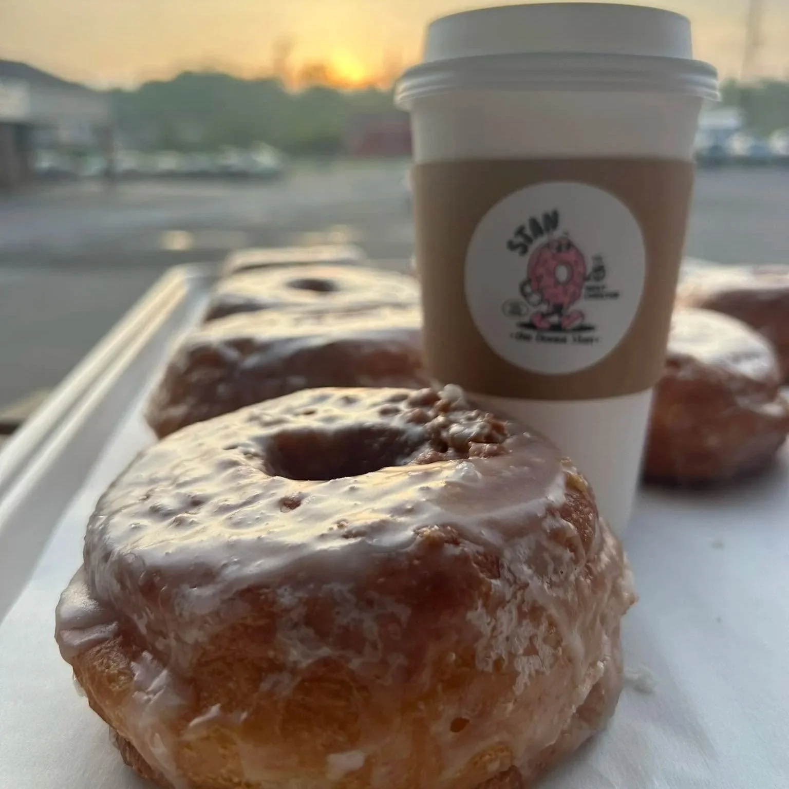 Close-up of glazed donuts and a coffee cup on a tray with a sunset in the background.
