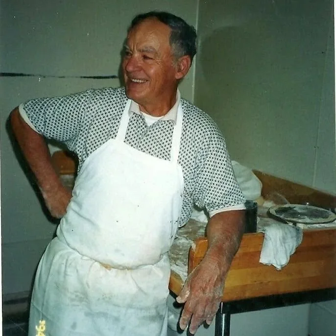 Our founder, Stan, smiling, wearing a white apron over a short-sleeved collared shirt with small patterned design, standing in a kitchen with a wooden counter and metal table, baking.