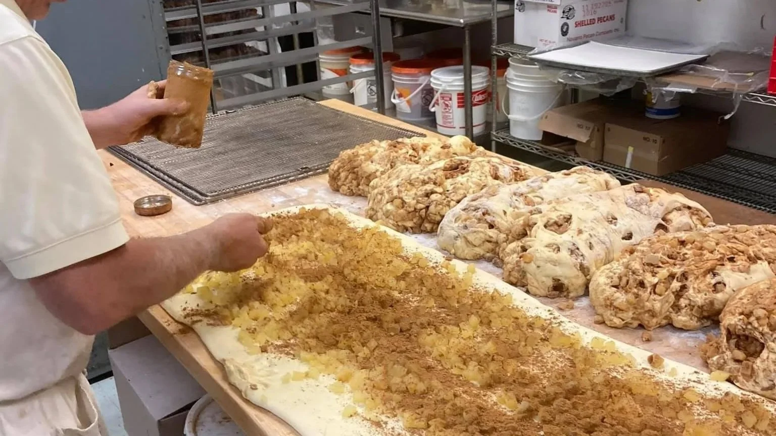 Bakery worker preparing fresh donuts with crumble topping on a large flat prep table.